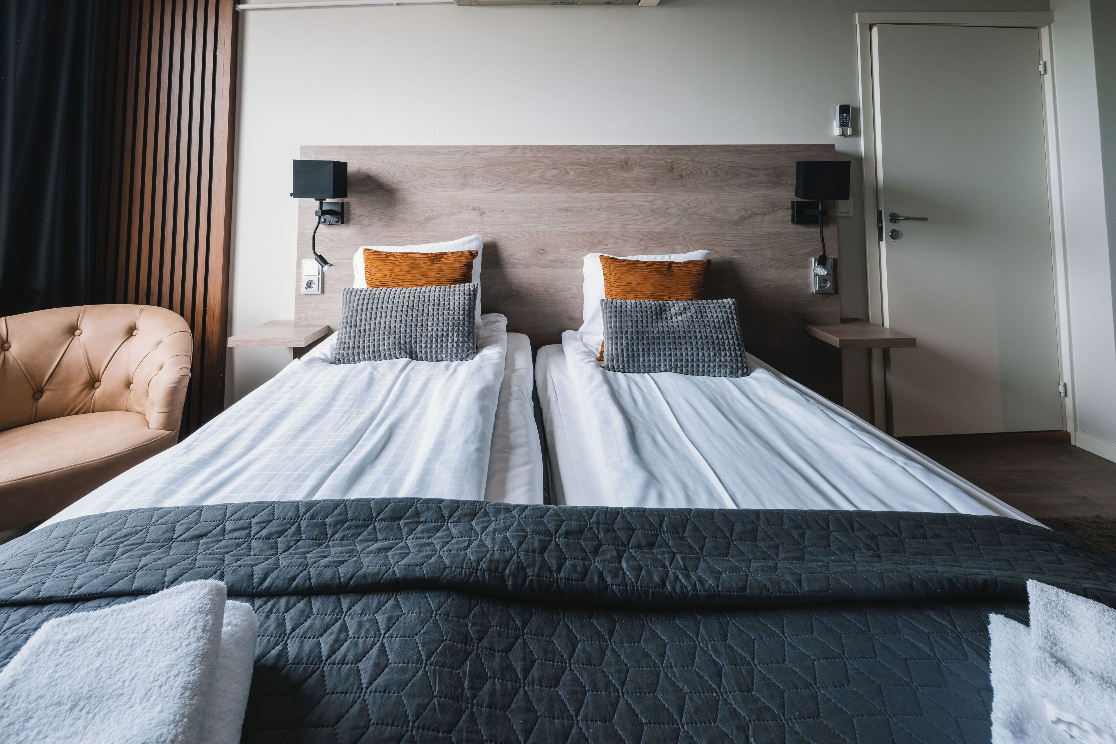 A hotel room with two beds, a wooden headboard, a bedside table with a lamp, and a chair.