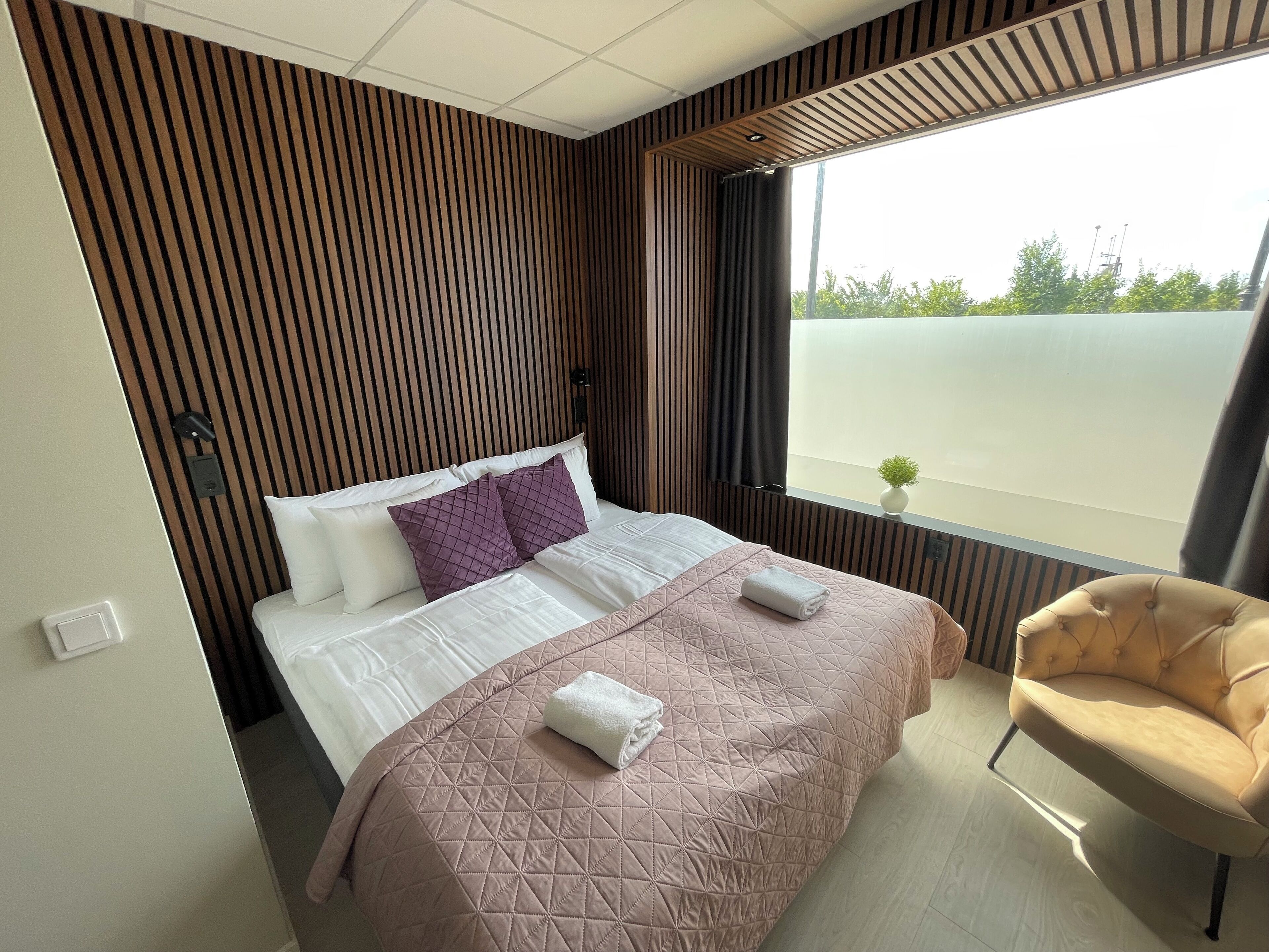 A modern hotel room with a large bed, a small side table, a chair, and a window with a view of greenery.