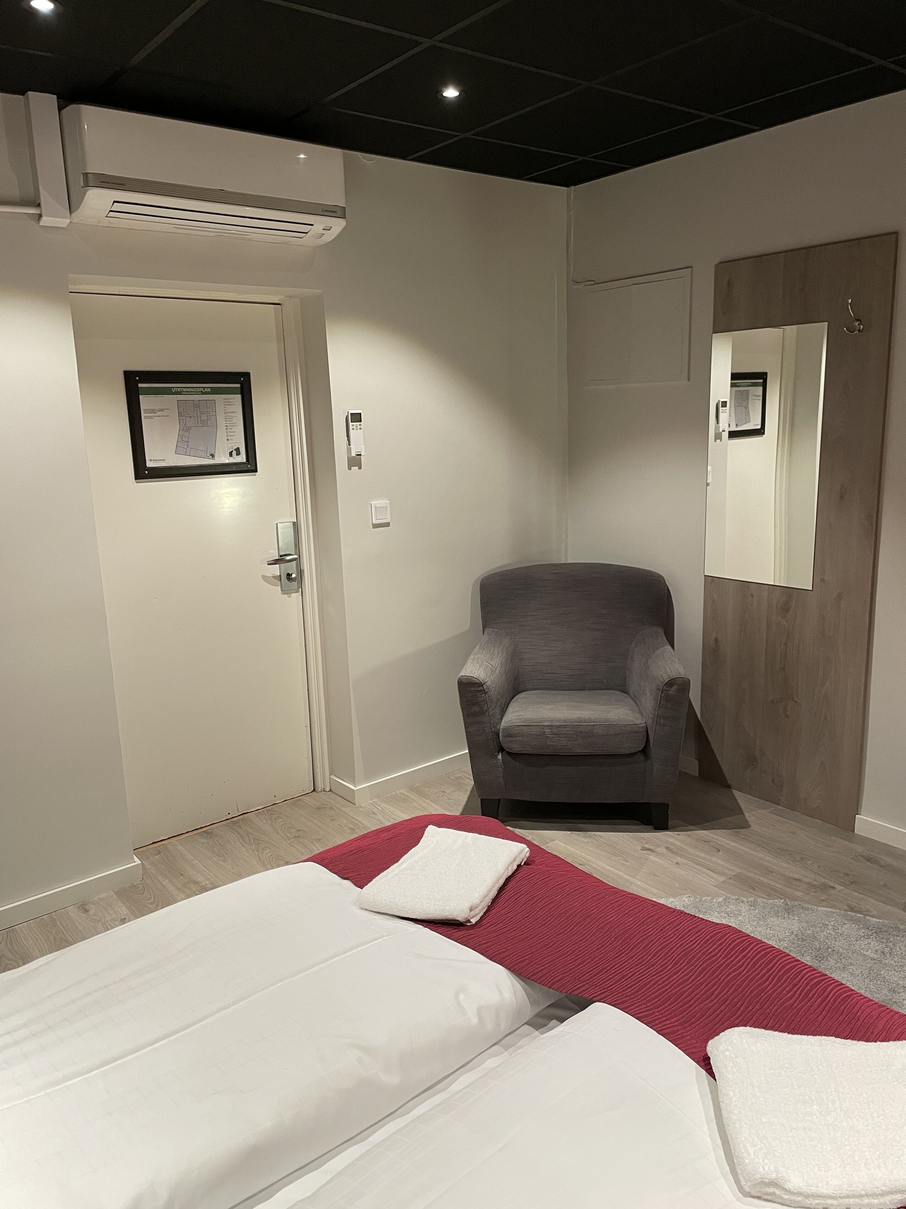 A hotel room with two beds, a gray armchair, a wooden door, and a mirror.