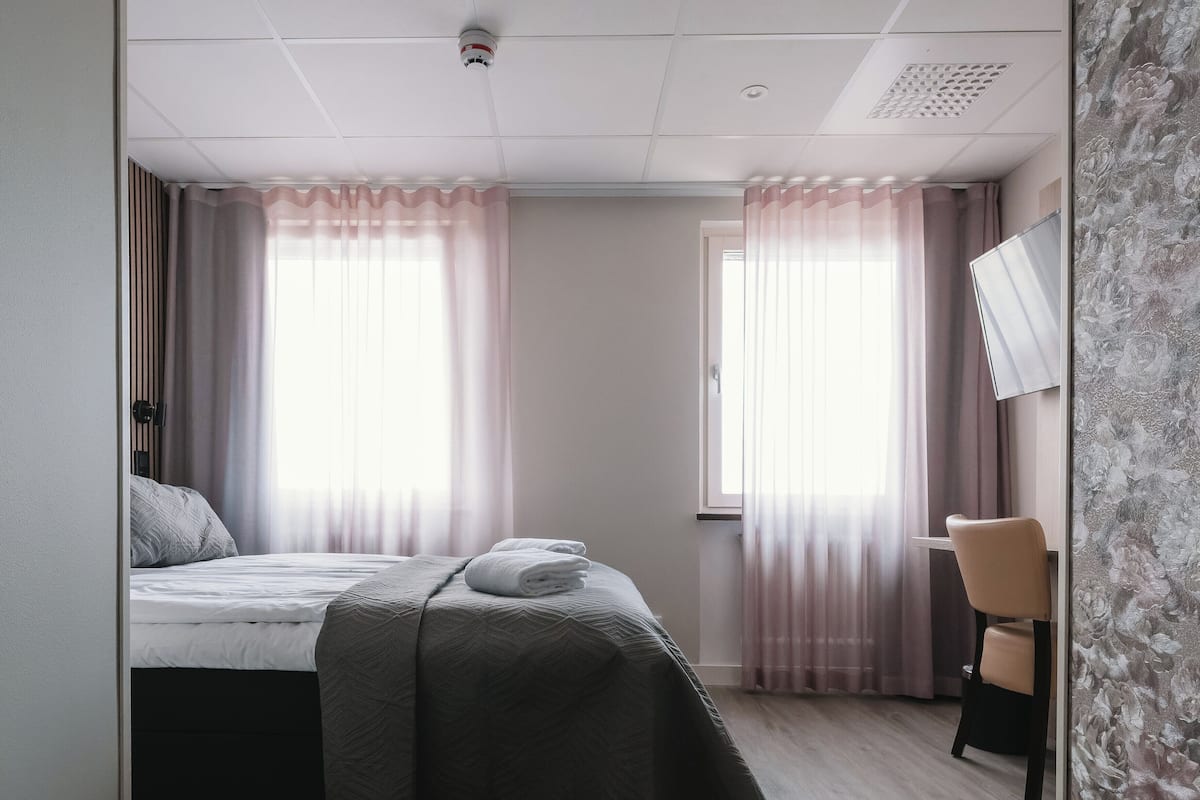 Habitación de hotel con cama, una silla y dos ventanas con cortinas.