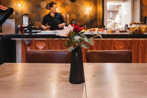 Interior de restaurant amb taula de fusta, un ram de flors i un taulell amb un cuiner al fons.