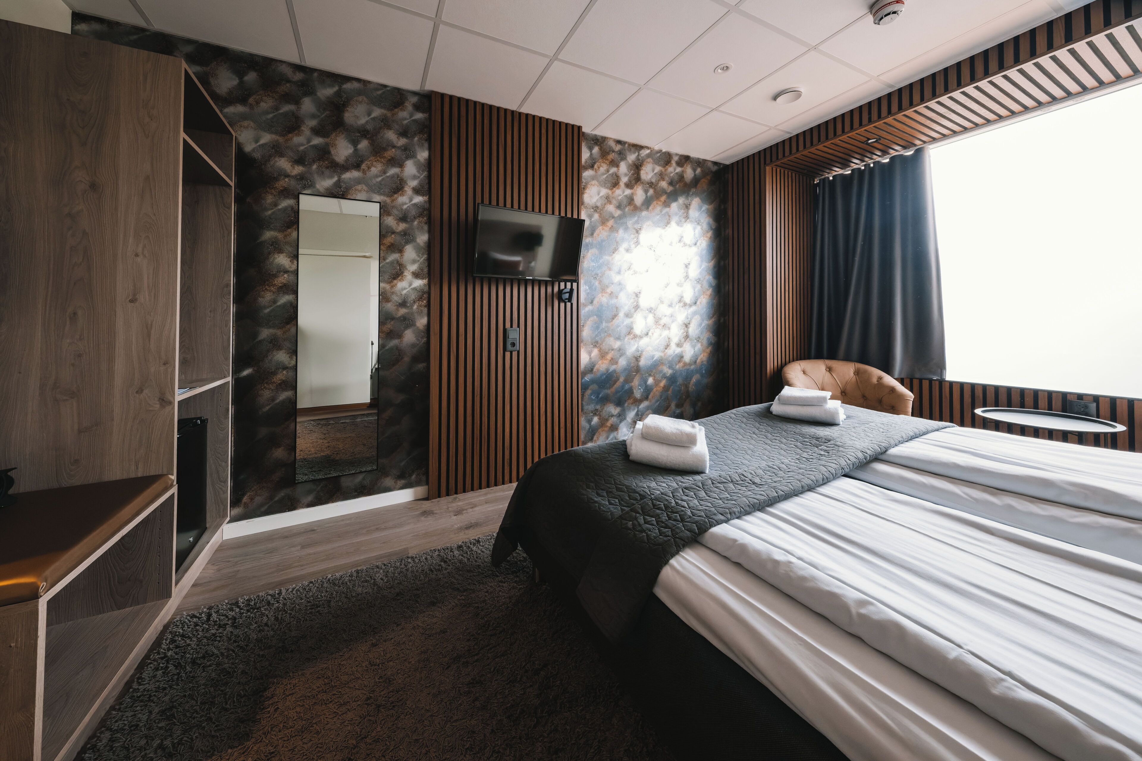 A hotel room with a bed, a TV, a wooden wardrobe, a mirror, and a window with curtains.