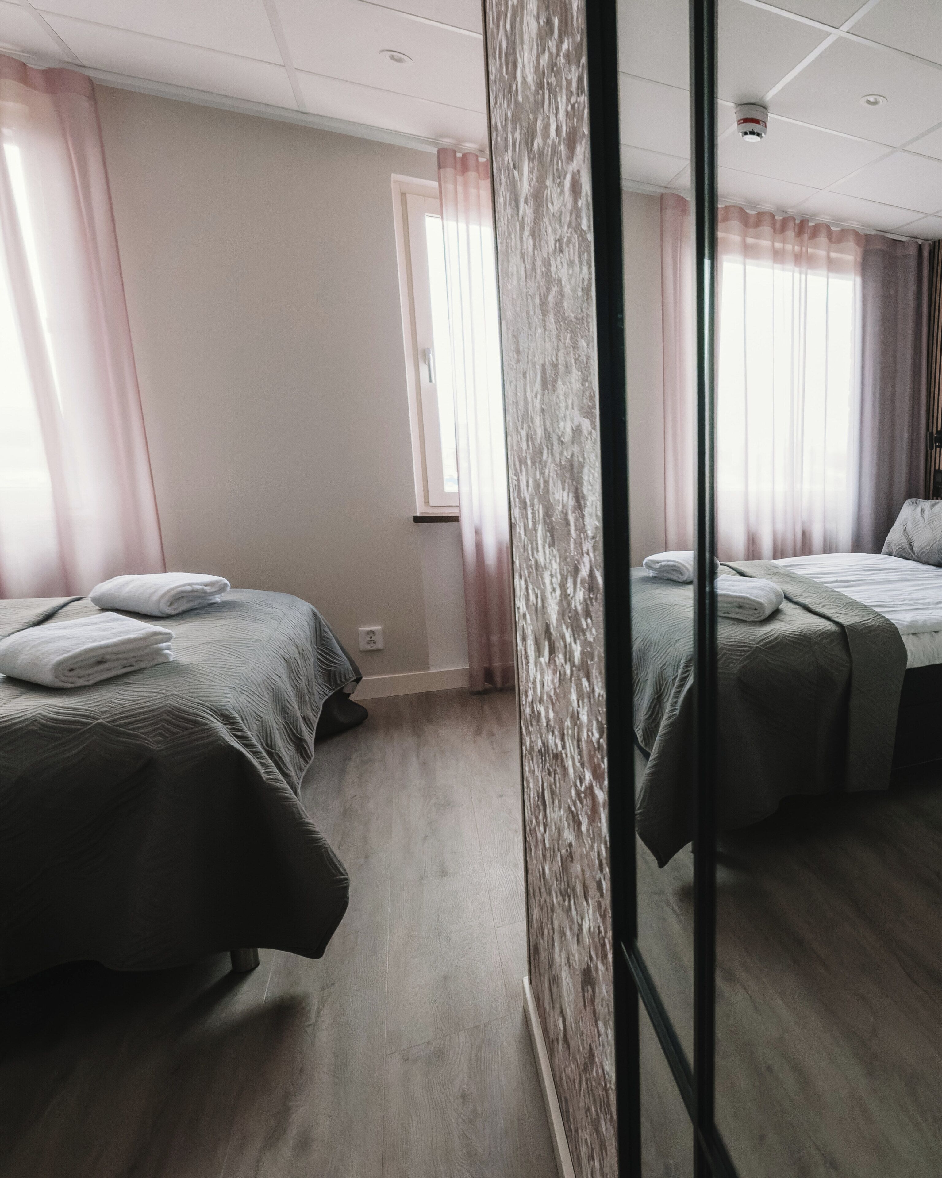 Ein Hotelzimmer mit zwei Betten, einem Fenster mit Vorhängen, Holzboden und einer sichtbaren Steckdose.