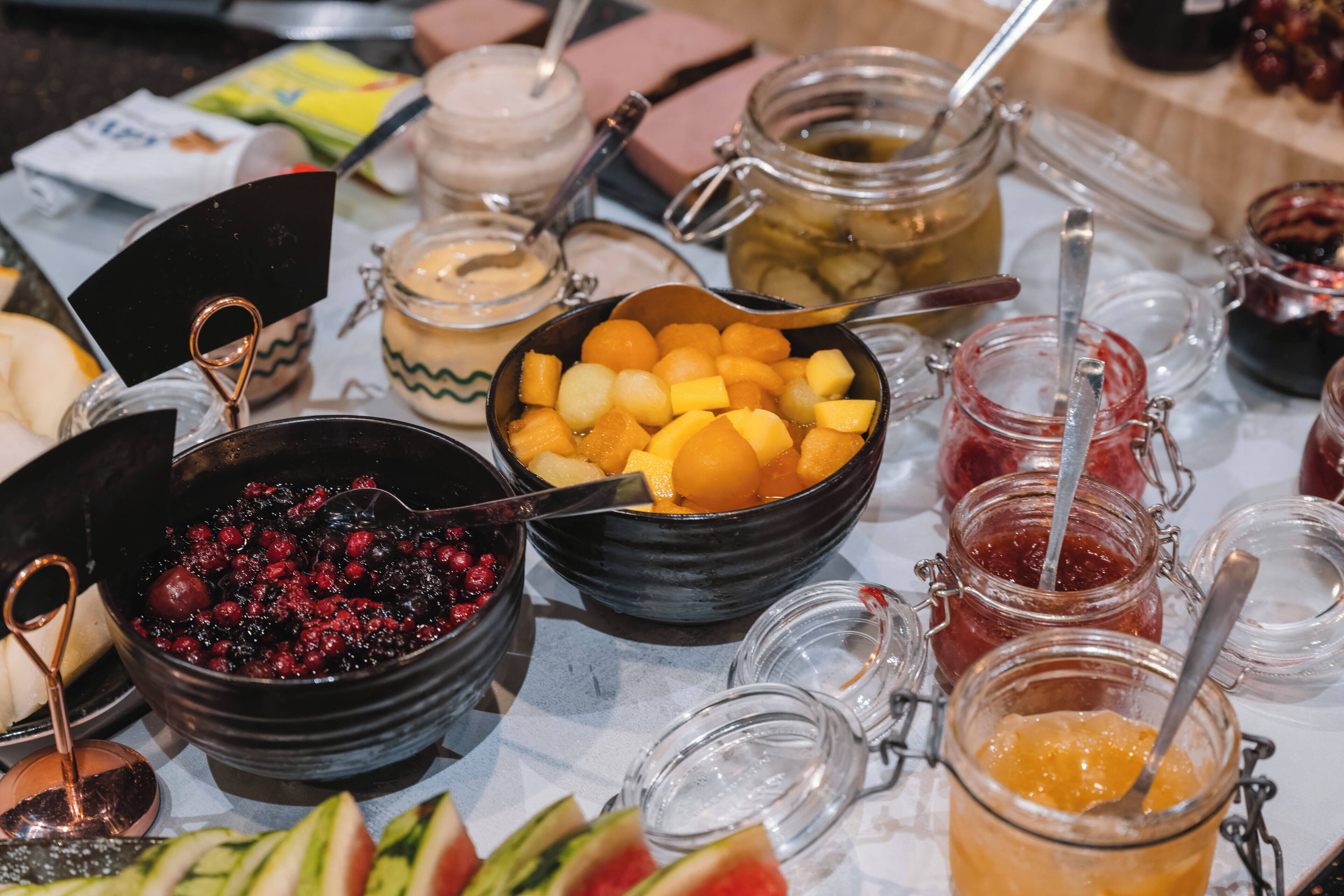 A table with various food items and condiments, including bowls of fruit, jars of preserves, and a metal spoon.