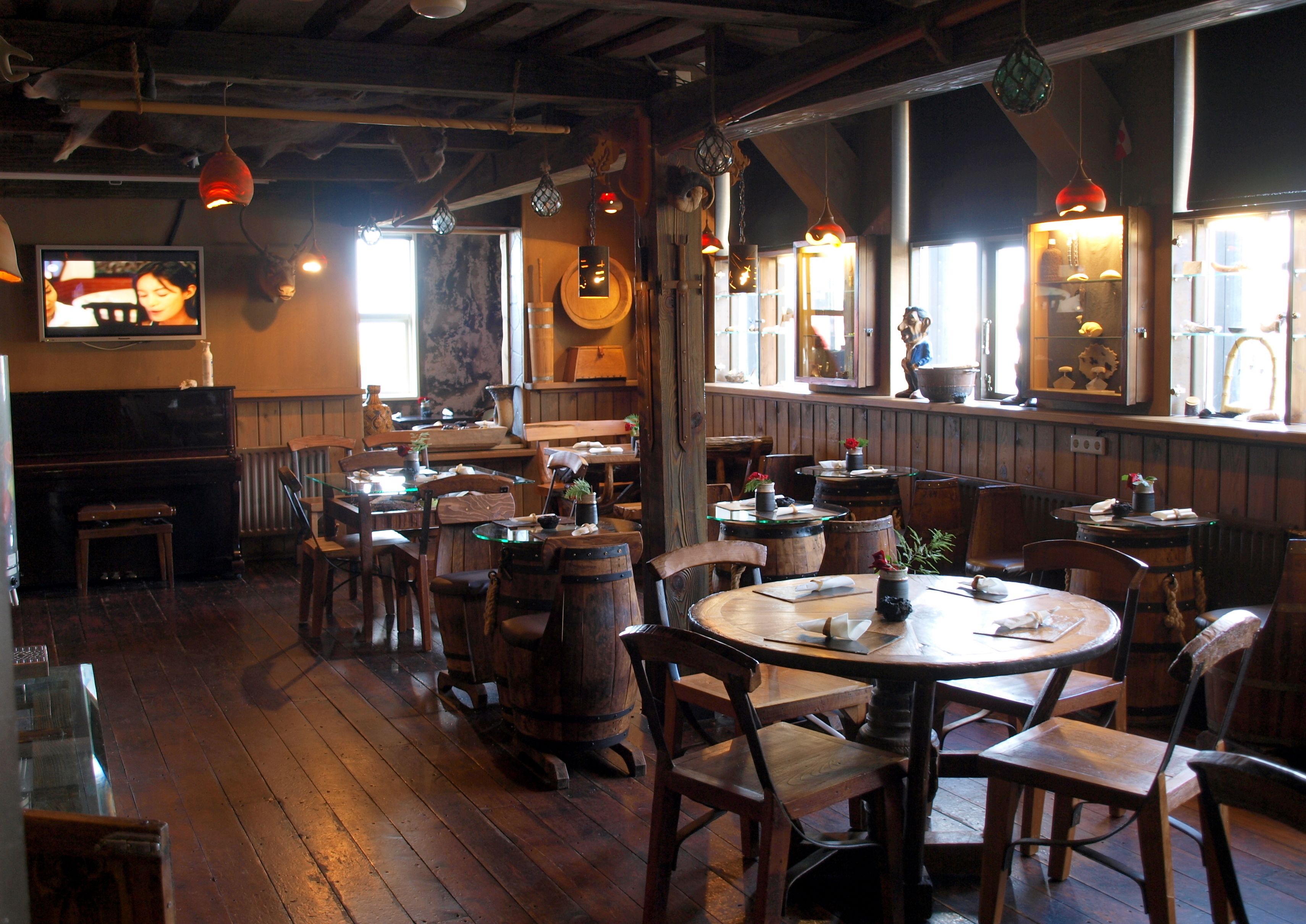 A rustic dining area with wooden tables and chairs, hanging light fixtures, and a television mounted on the wall.