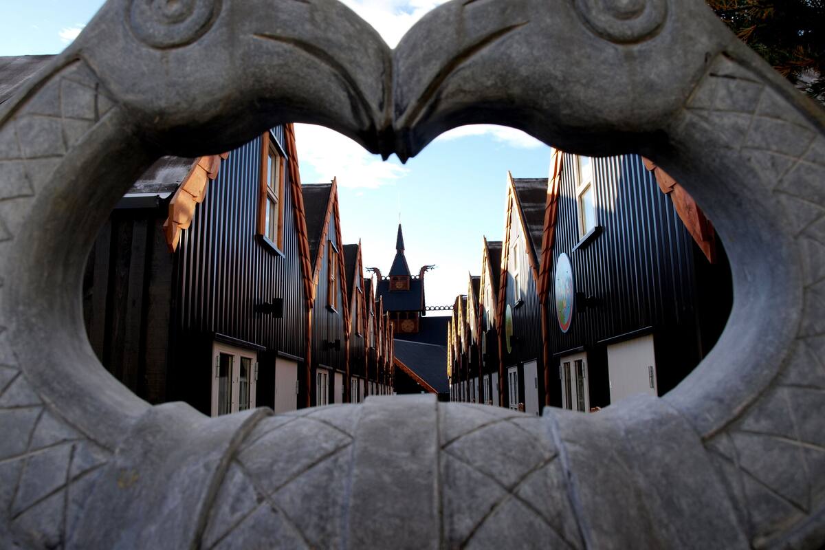 A stone archway with a heart-shaped design, leading to a row of traditional buildings with wooden facades and a clock tower in the background.