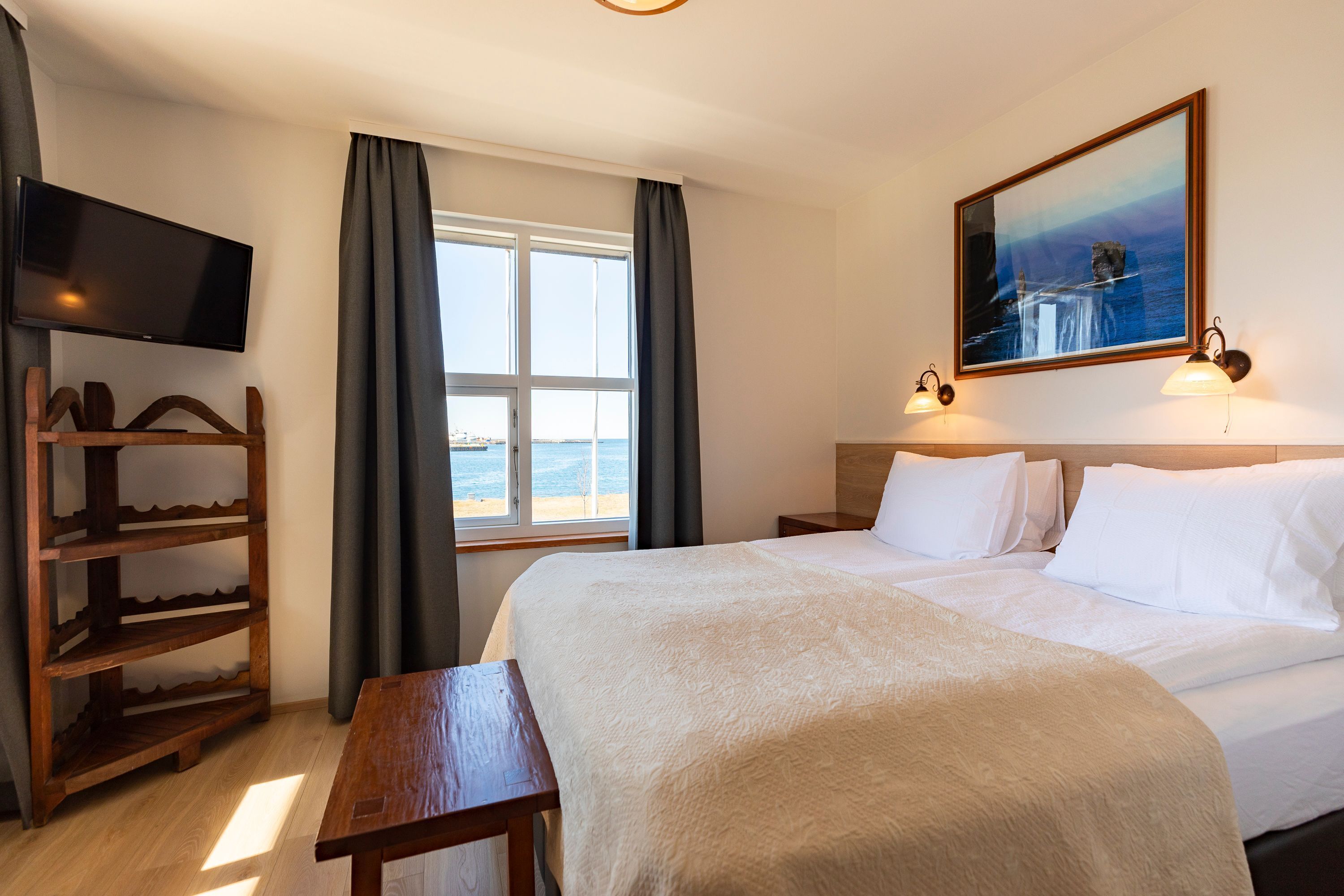 A hotel room with a bed, a TV, a wooden shelf, a window with curtains, and a painting on the wall.