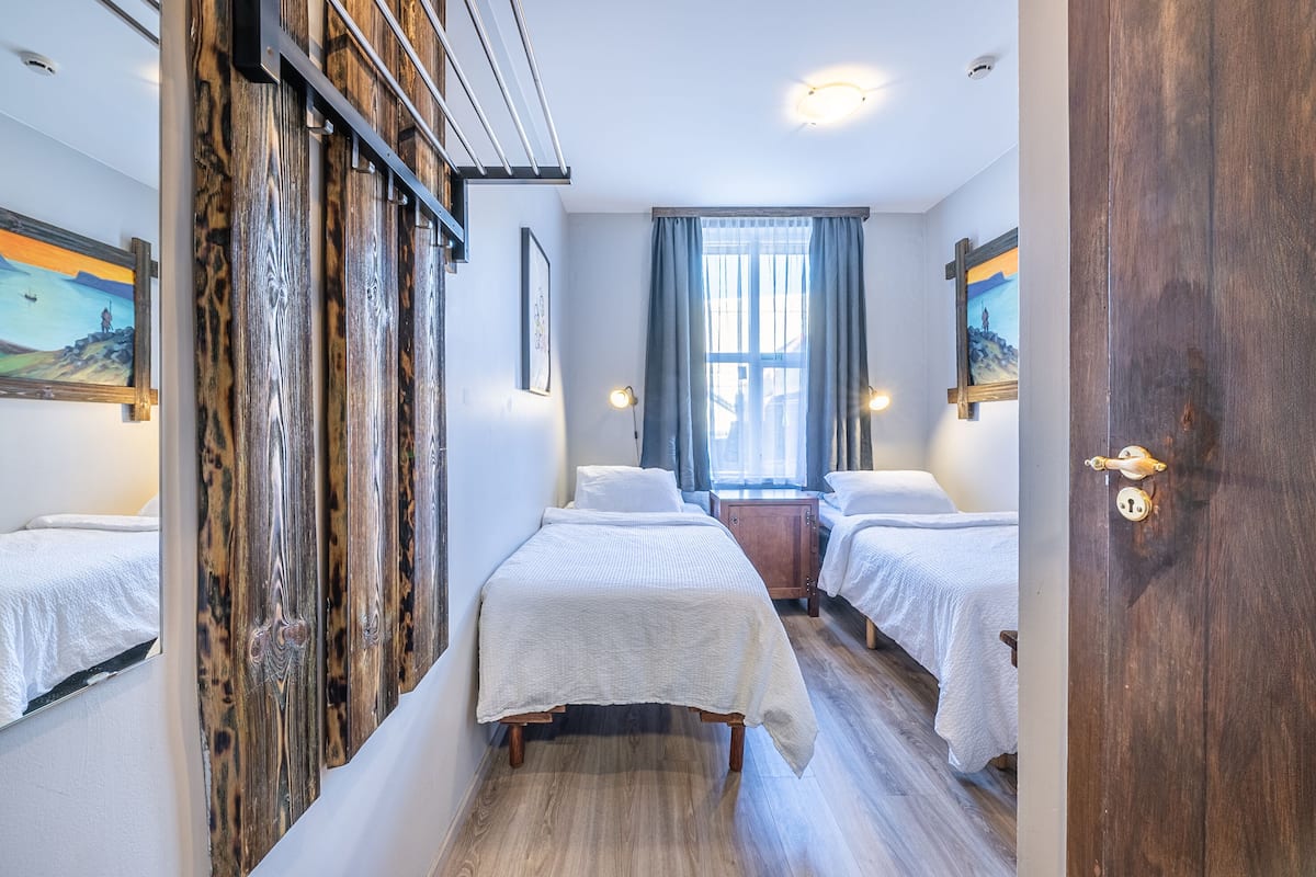 A hotel room with two beds, a wooden wardrobe, a window with curtains, and artwork on the walls.