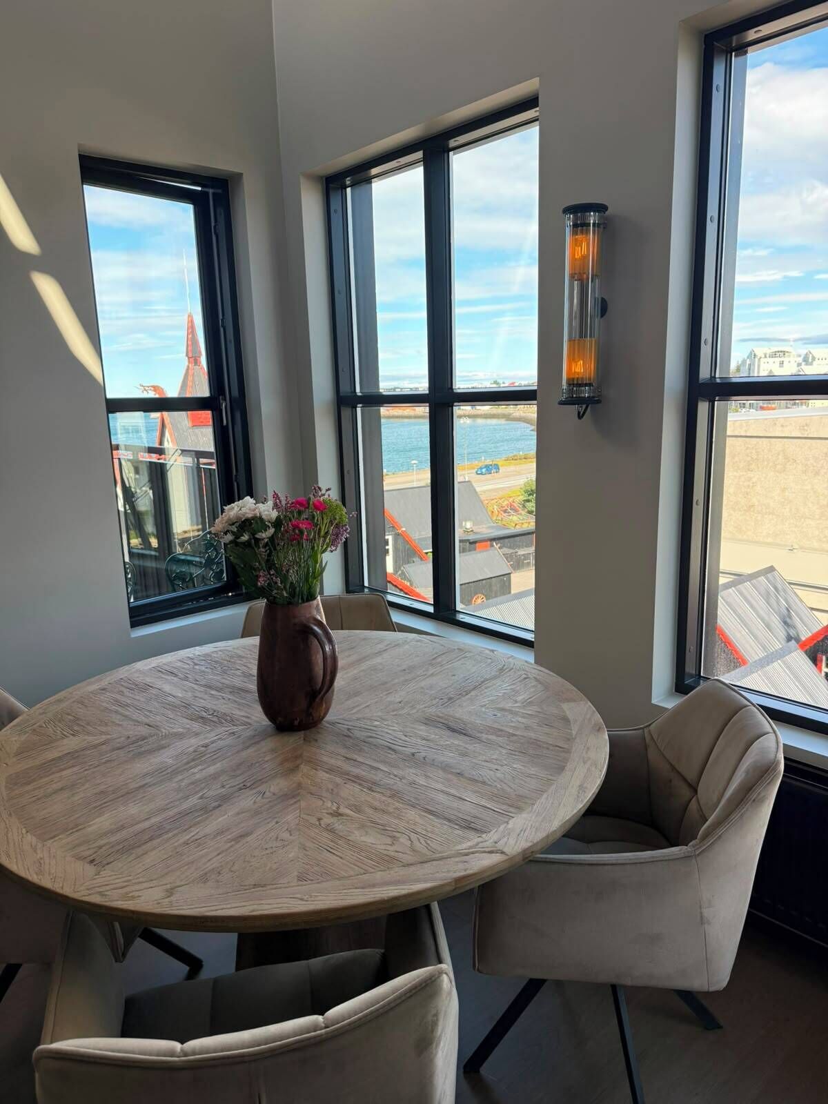 Ein runder Esstisch mit Blumen in einer Vase, zwei Stühle und große Fenster mit Blick auf Gebäude und ein Gewässer.