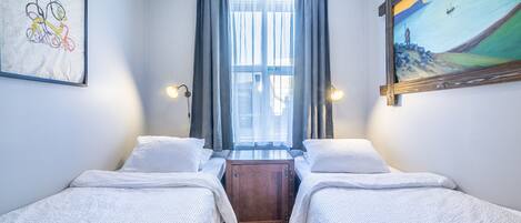 A room with two single beds, a wooden nightstand, a window with curtains, wall art, and two wall-mounted lights.