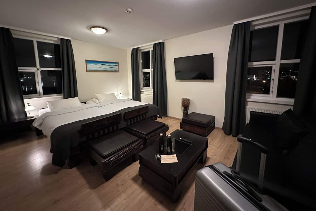 A hotel room with a large bed, a sofa, a small table, a TV, and a window with a city view.
