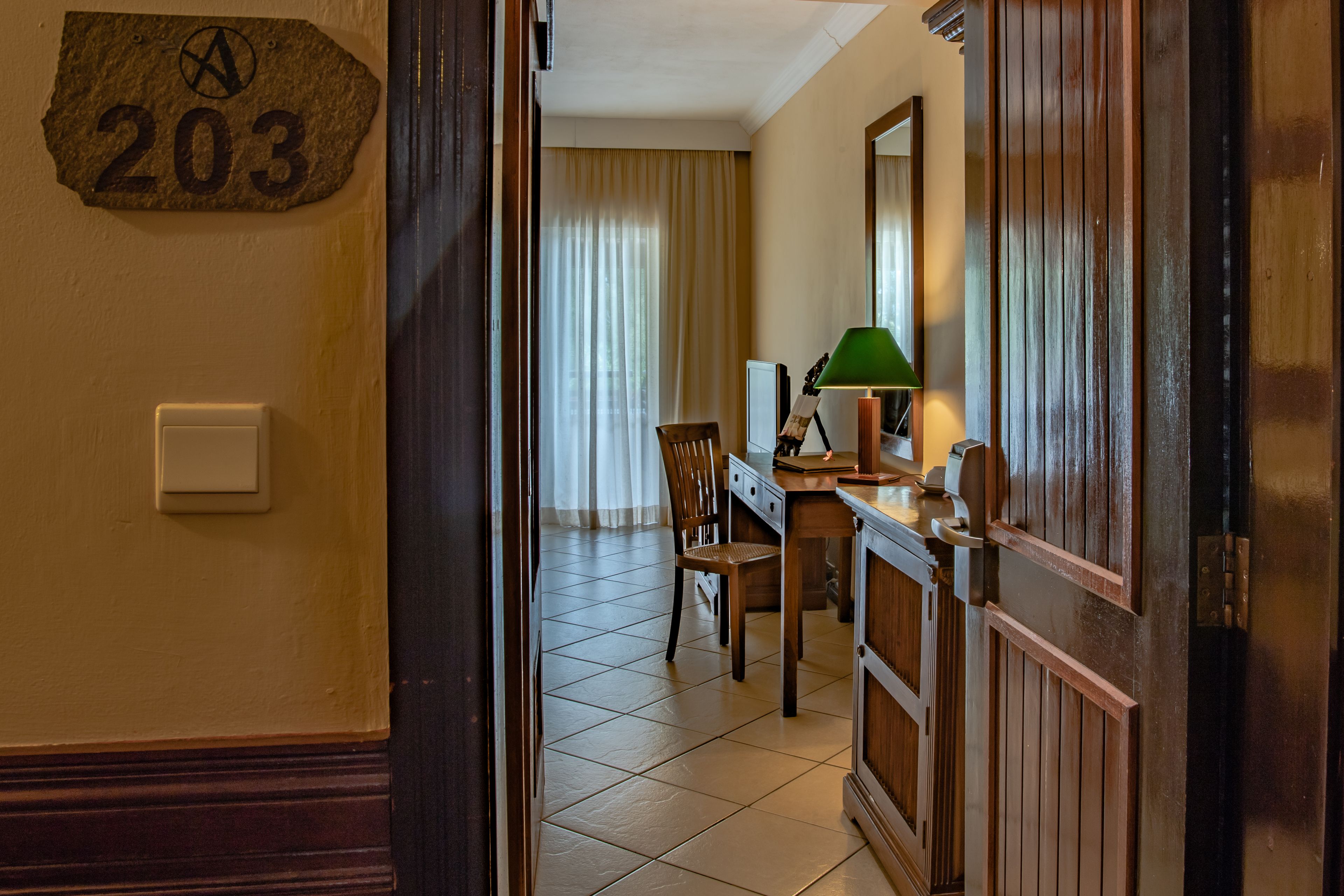 Une chambre d’hôtel avec une porte en bois, une plaque portant le numéro « 203 », un bureau avec une lampe, une chaise et une fenêtre avec des rideaux.