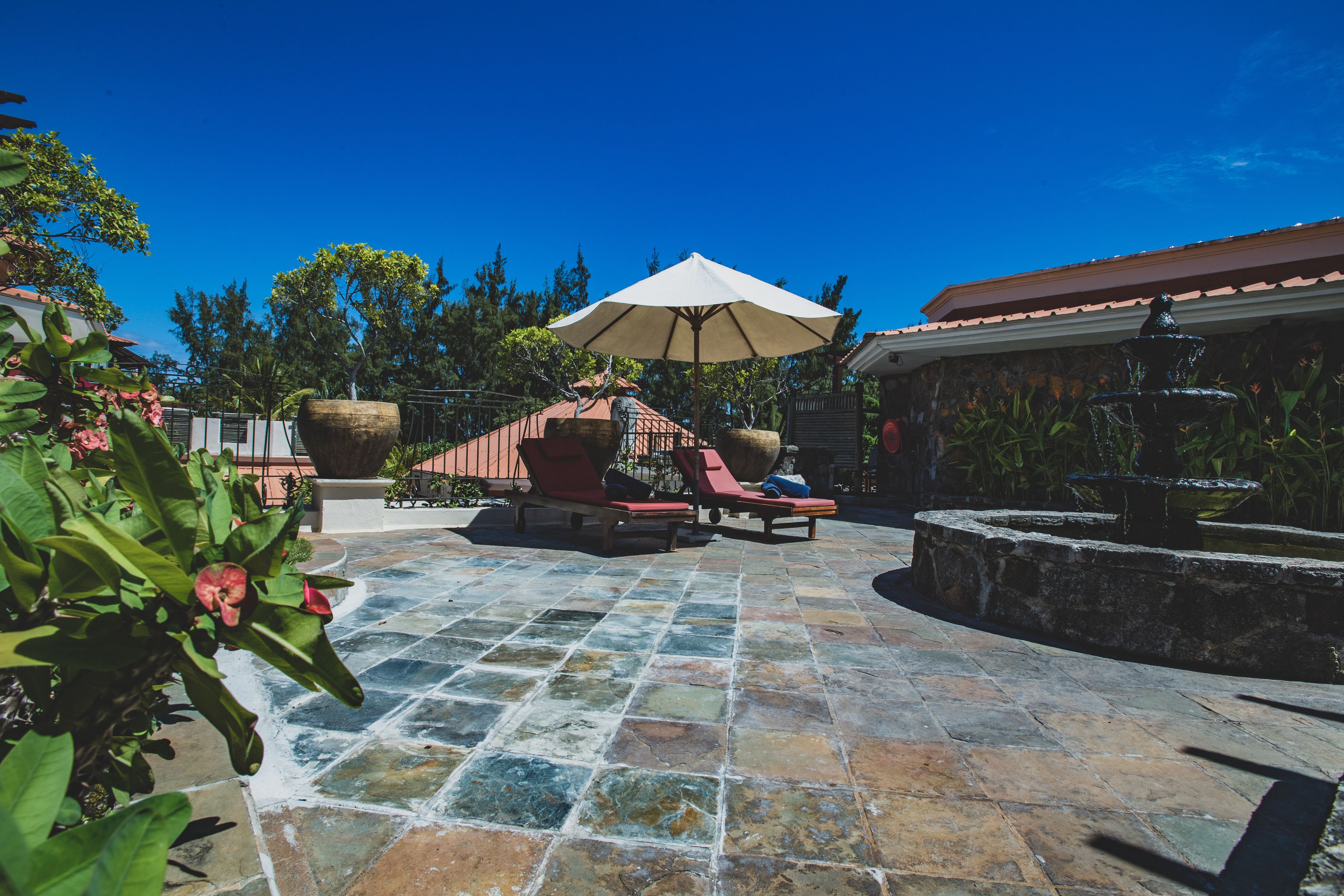 Un patio con suelo de piedra, dos sillones de descanso, una sombrilla grande y una fuente.