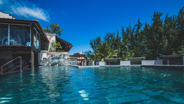 A clear swimming pool with a waterfall feature, surrounded by a building with large windows and a balcony. - Aanari Hotel and Spa (Flic-en-Flac)