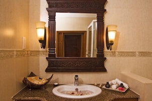 Un baño con un espejo grande, dos luces empotradas, un lavamanos con un recipiente decorativo y una bandeja con toallas y artículos de aseo.
