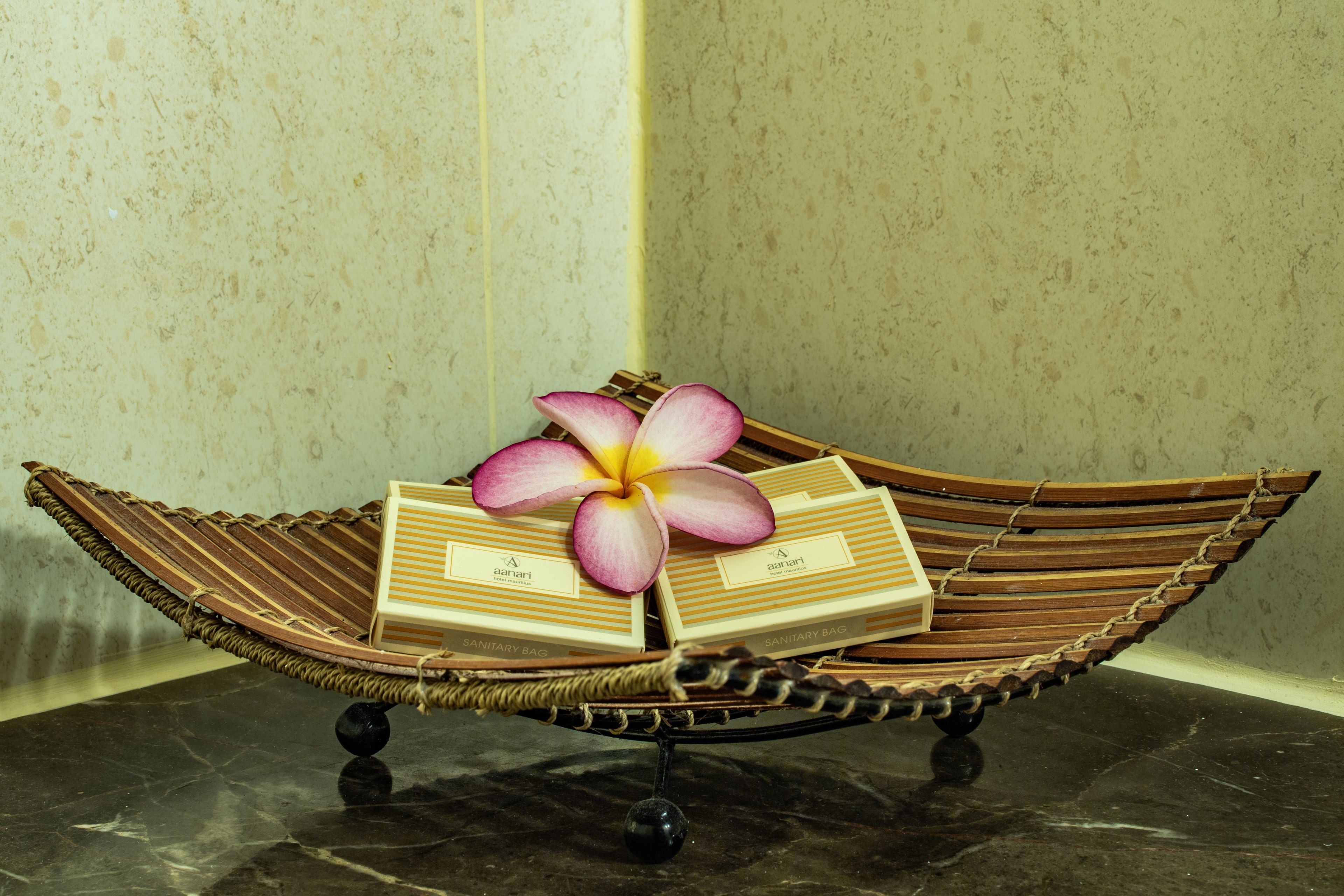 Una silla tejida con forma de barco, con base negra, que sostiene tres bolsas sanitarias y una flor rosa y amarilla.