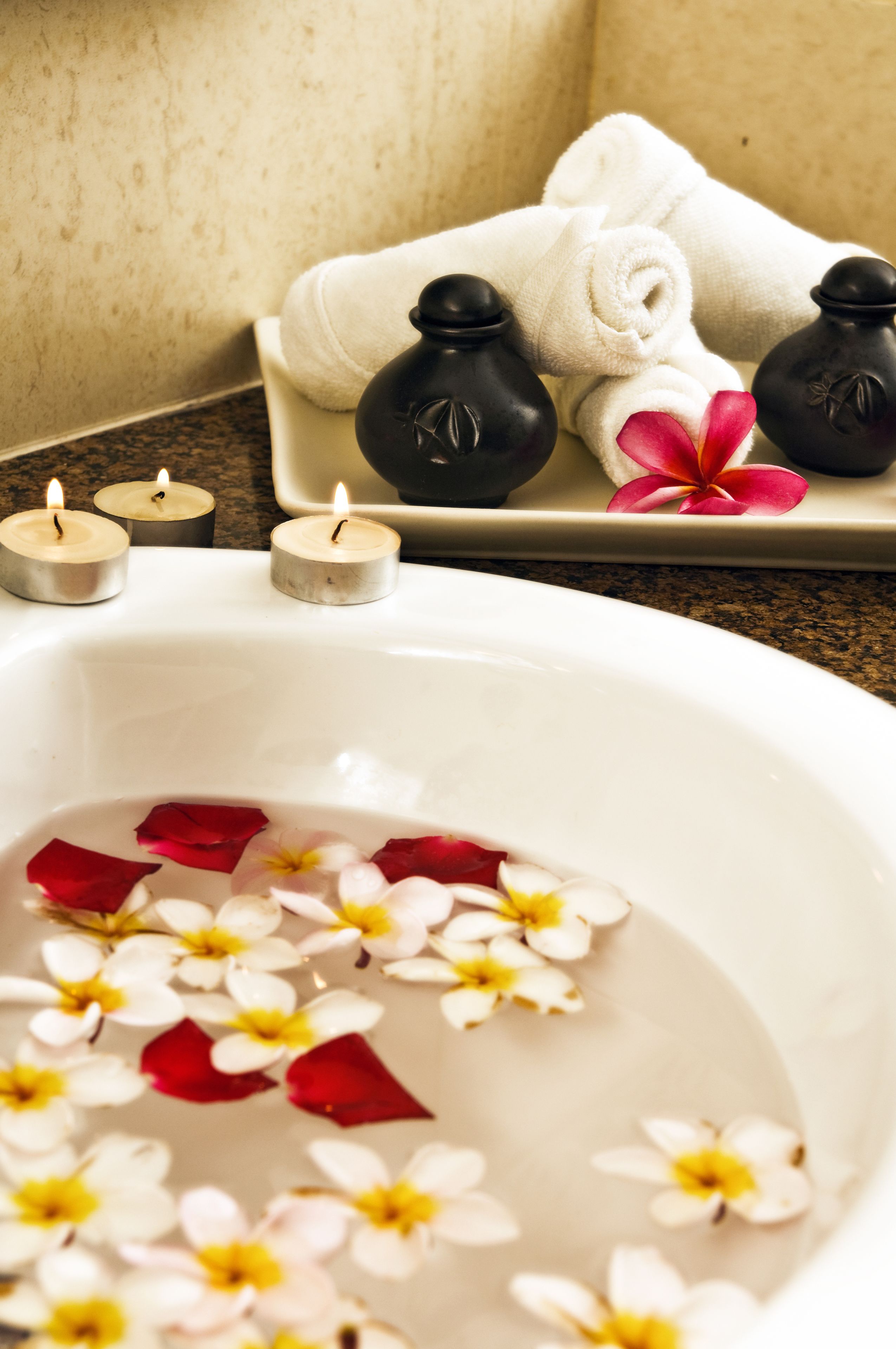 A spa setting with a bathtub filled with water and flower petals, a white towel, rolled towels, and black massage stones on a tray.