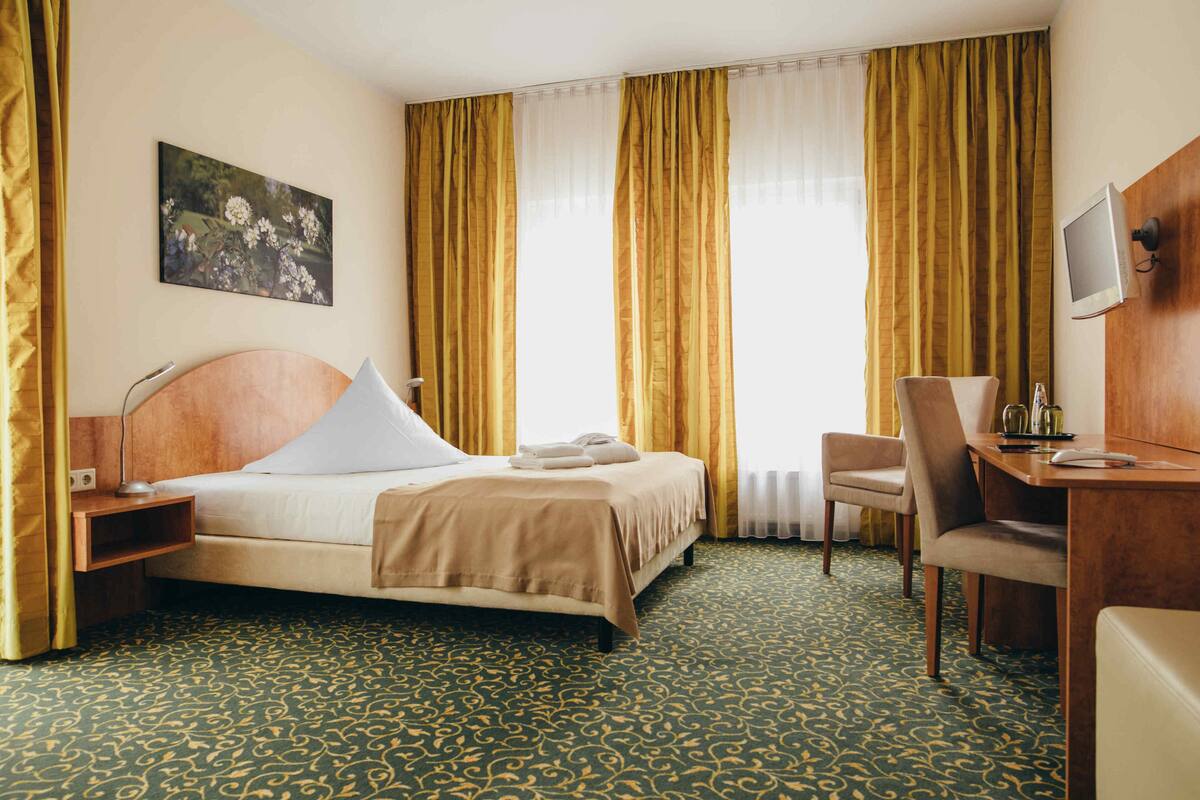 A hotel room with a bed, a desk, a chair, a television, and a painting on the wall.
