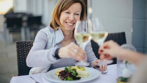 Una persona sentada en una mesa al aire libre con un plato de comida y una copa de vino blanco.