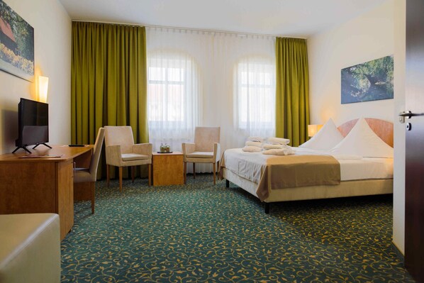 A hotel room with a bed, two chairs, a desk, a lamp, and a painting on the wall.