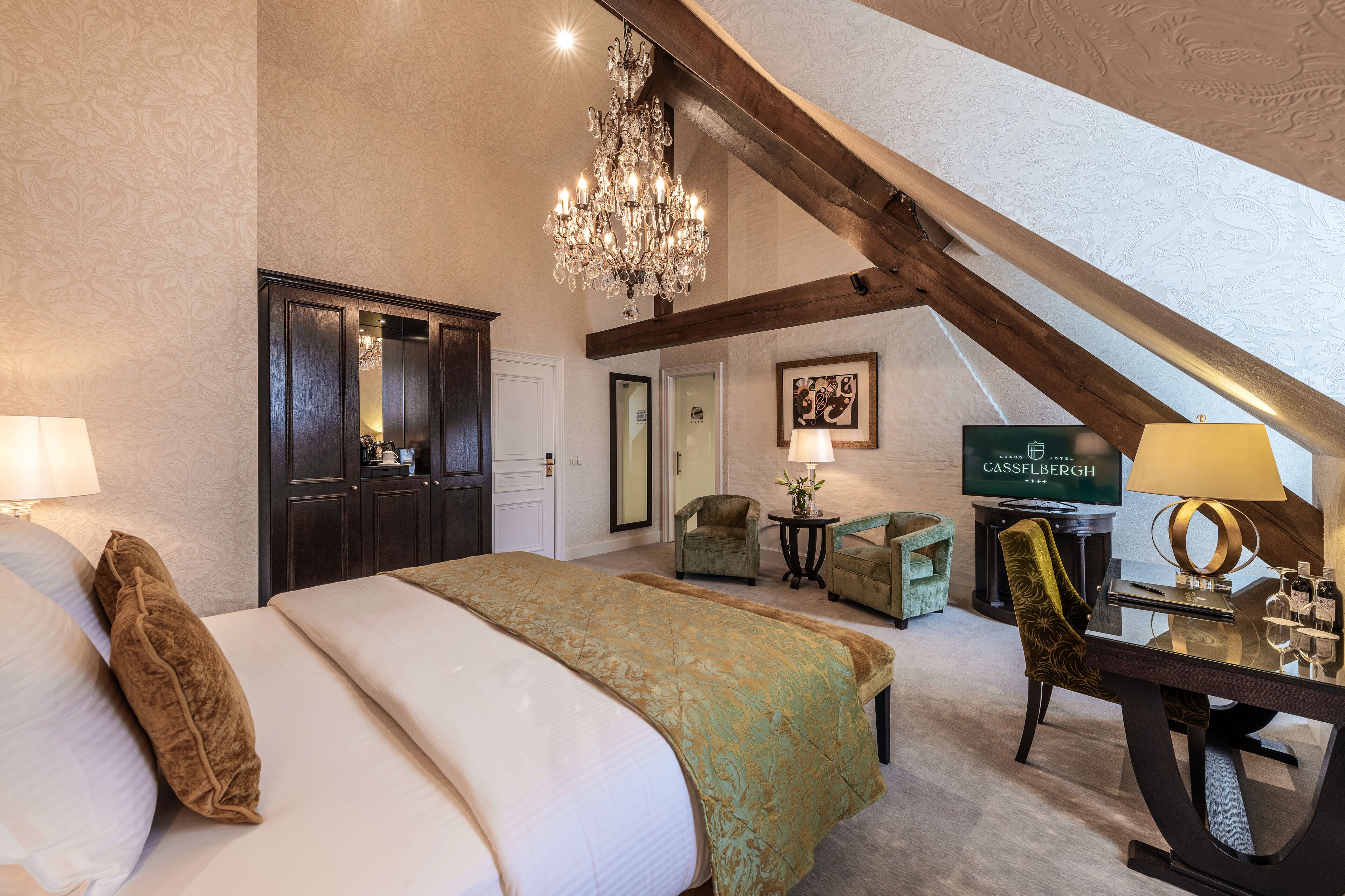 Una habitación de hotel con una cama grande, un candelabro, un escritorio con una lámpara y un televisor mostrando el nombre del hotel.