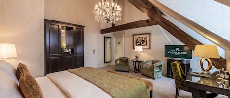 Una habitación de hotel con una cama grande, un candelabro, un escritorio con una lámpara y un televisor mostrando el nombre del hotel.