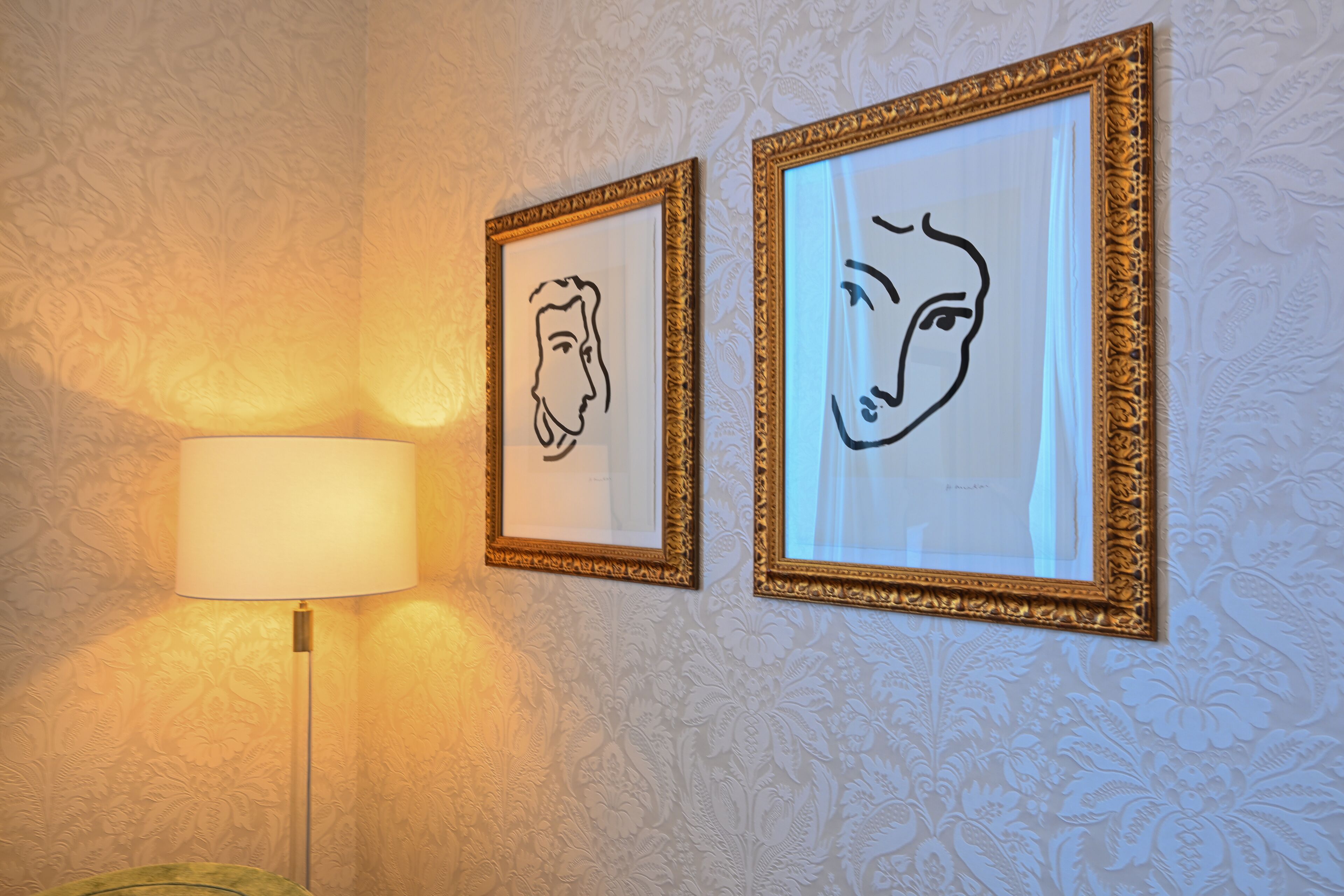 Two framed line drawings on a wall with a patterned wallpaper background and a floor lamp to the left.