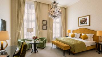 A hotel room with a bed, two chairs, a small table, a bench, a chandelier, and a painting on the wall.