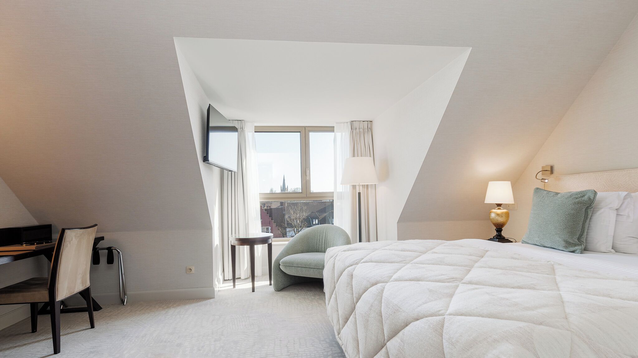 A hotel room with a bed, a desk, a chair, a lamp, and a view of a cityscape through a window.