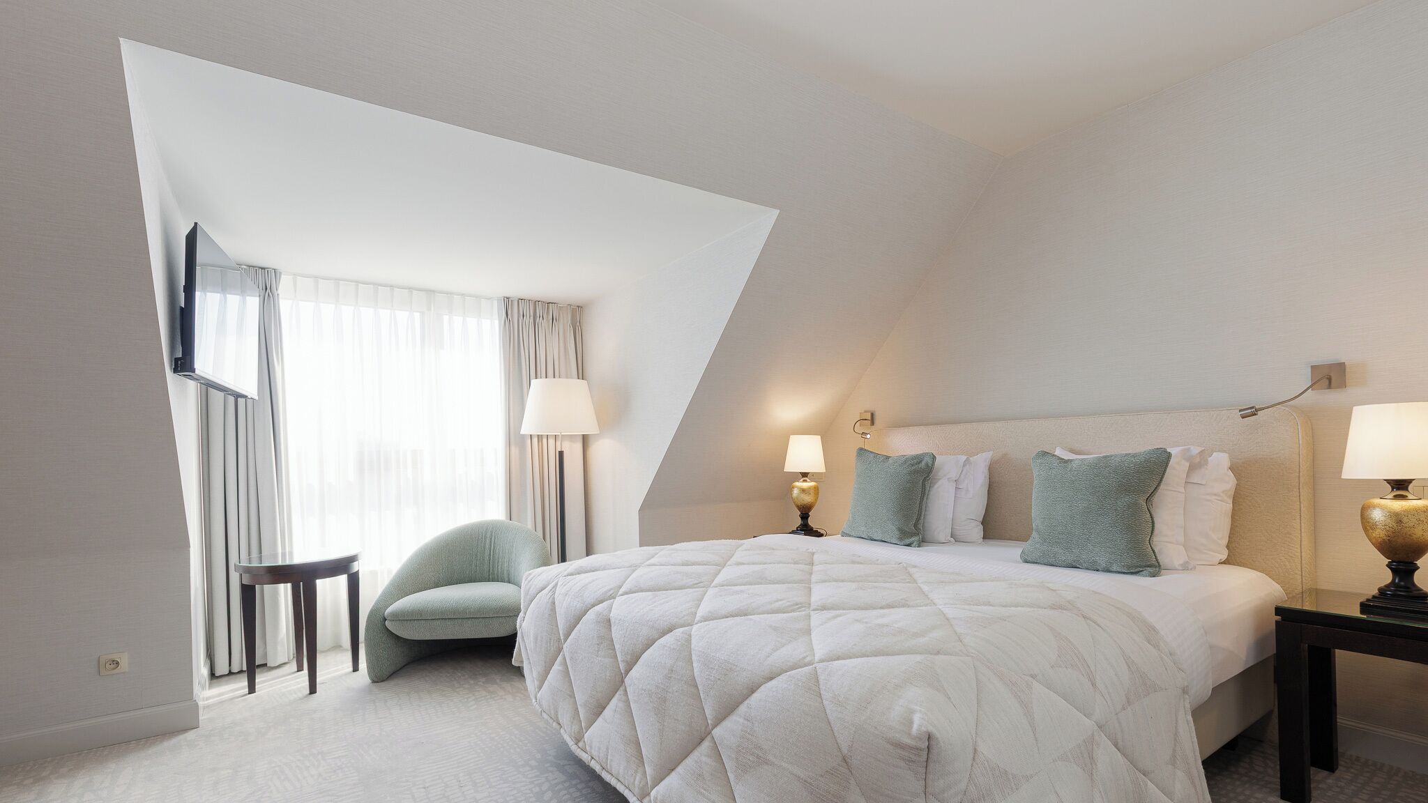 Una habitación de hotel moderna con una cama grande, dos mesitas de noche, una silla y una ventana con cortinas.