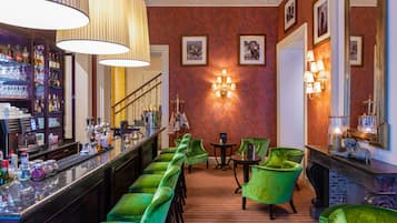 Una zona de bar con sillas de terciopelo verde, una barra con diferentes botellas y una lámpara fijada a la pared.