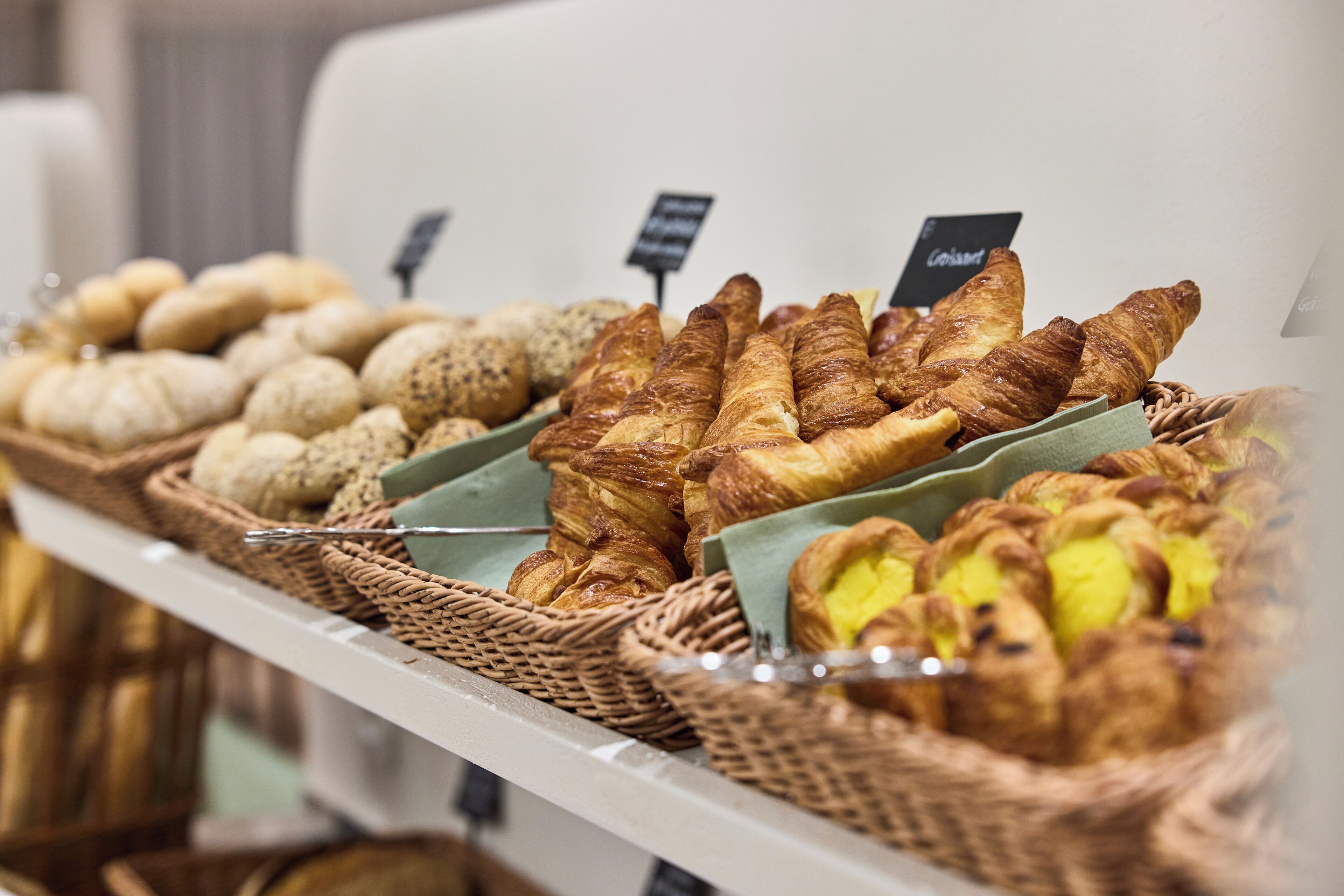 Mostrador de panadería con diferentes tipos de pan y pasteles, incluyendo croissants y panecillos, dispuestos en estantes.