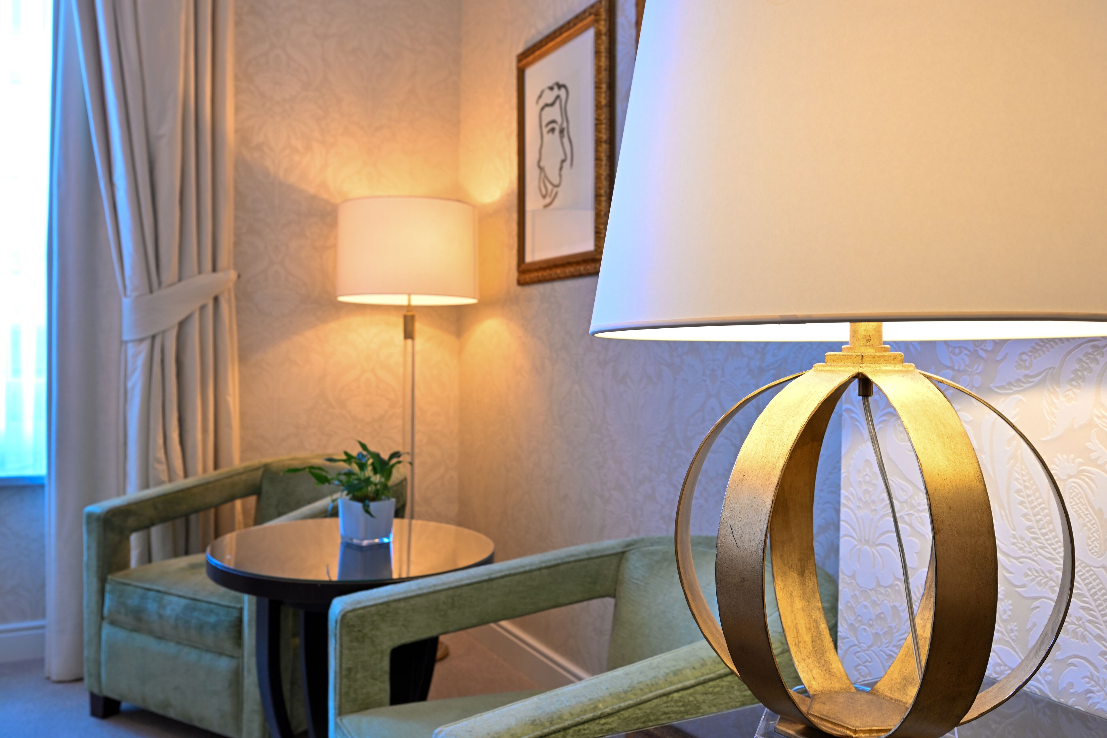 A room with a green armchair, a round table with a potted plant, a wall-mounted lamp, and a framed picture.