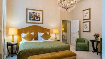 A hotel room with a large bed, two bedside tables with lamps, a green armchair, a bench, a chandelier, and artwork on the walls.