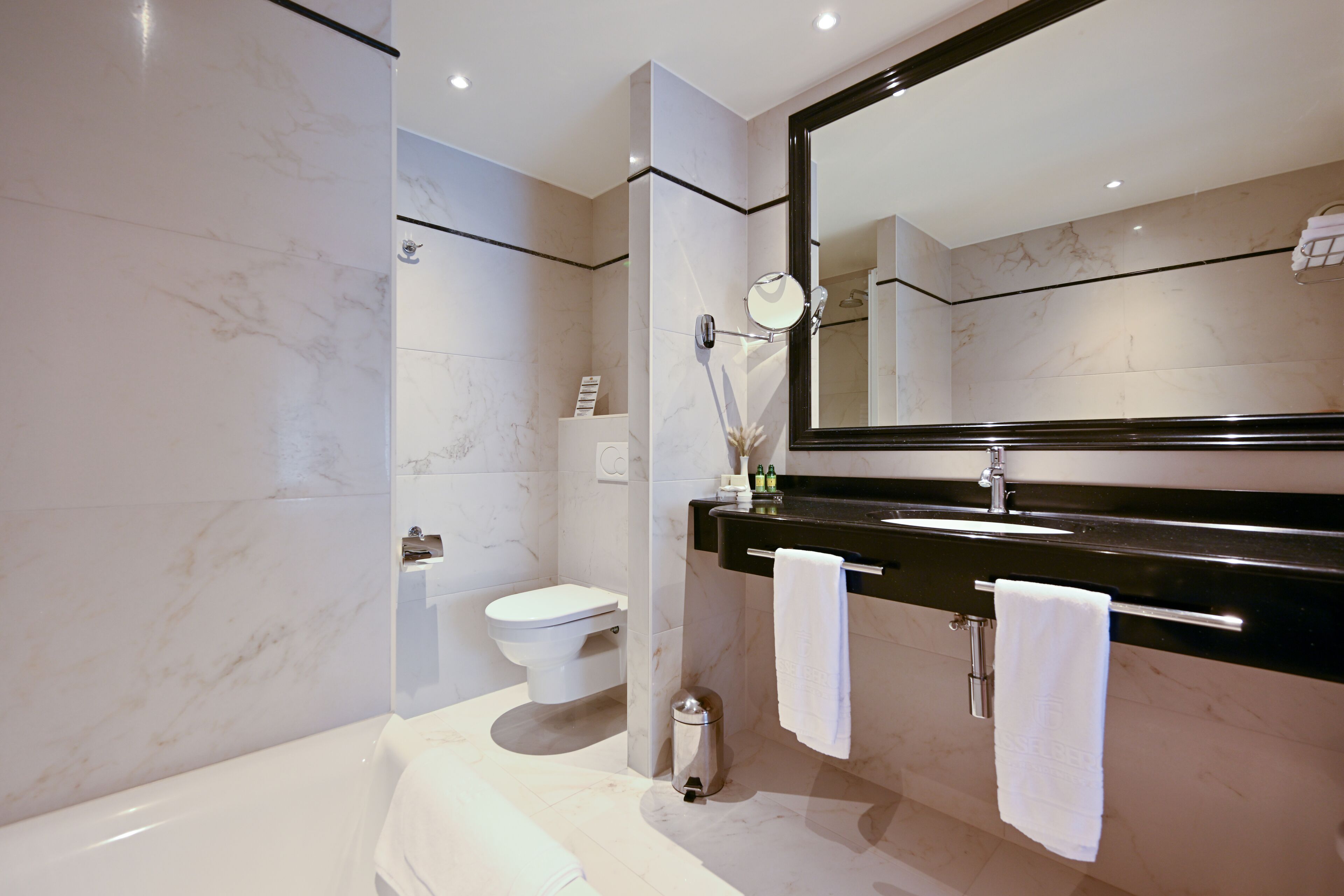 Un baño moderno con inodoro blanco, bañera, un espejo grande y una encimera negra con toallero.
