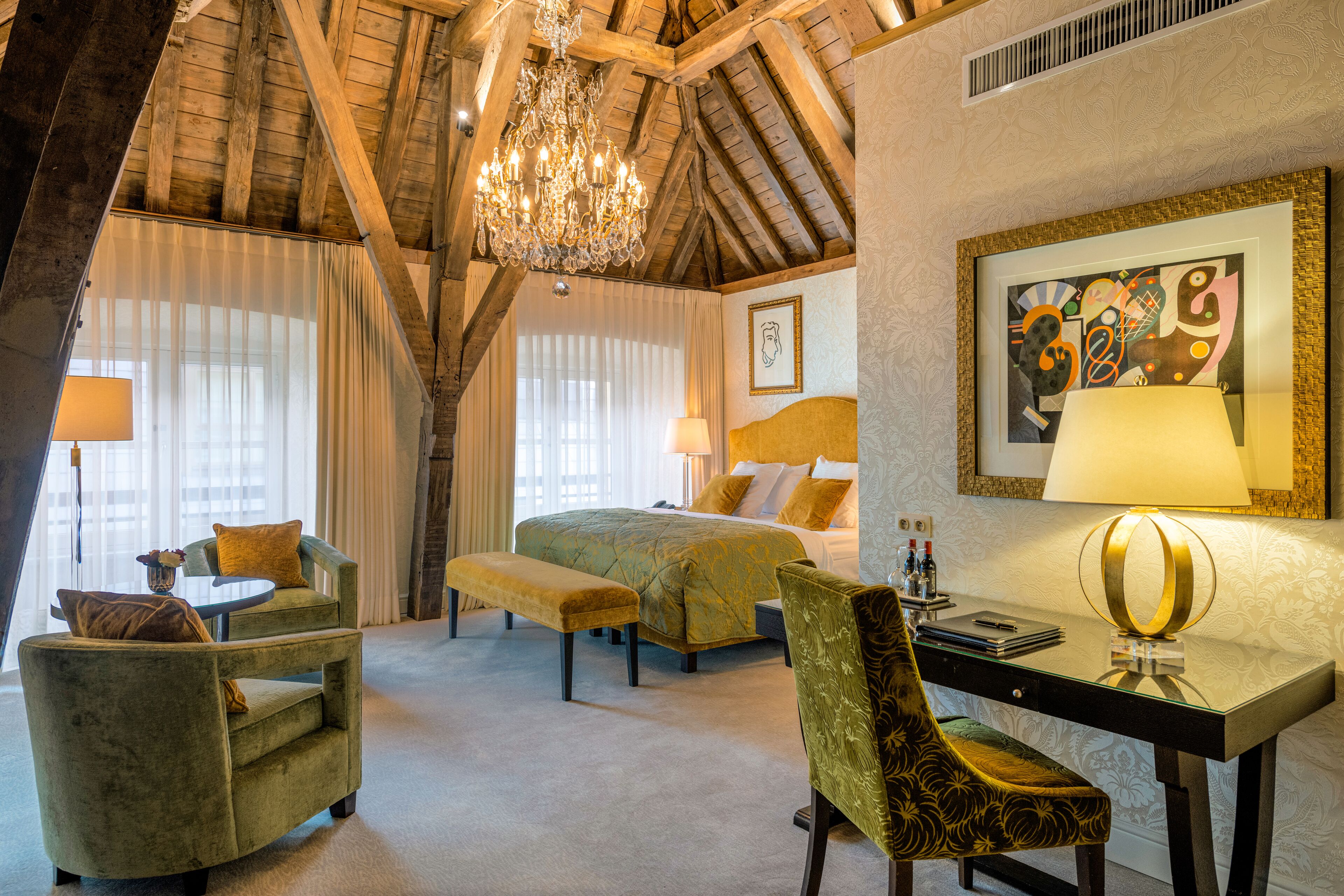 Quarto de hotel com uma cama, duas poltronas, uma escrivaninha e um lustre.