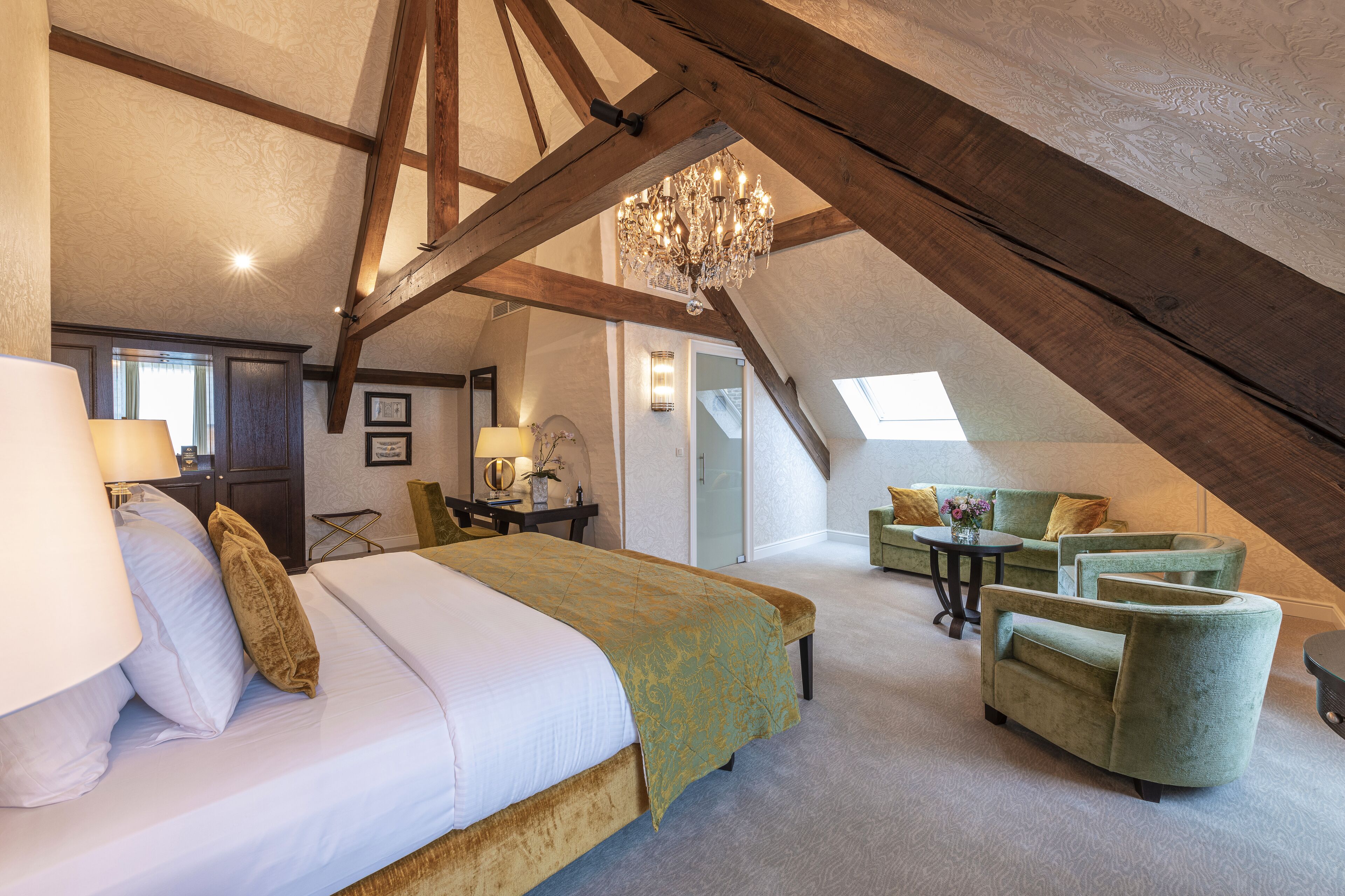 A spacious attic bedroom with a large bed, a chandelier, and a seating area with armchairs.