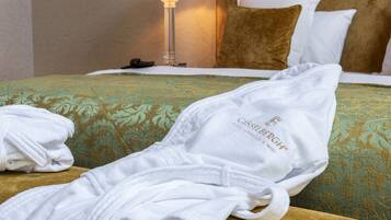 Una cama bien tendida con una colcha verde estampada, almohadas blancas y una bata de baño blanca con texto.