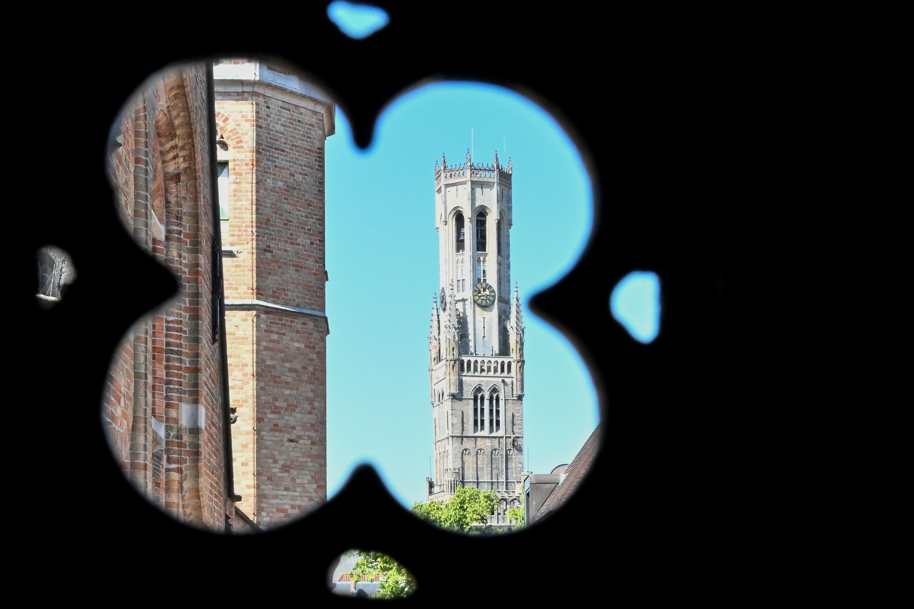 A view through a circular window of a tall brick tower with a clock, a clear blue sky, and a small tree visible in the foreground.