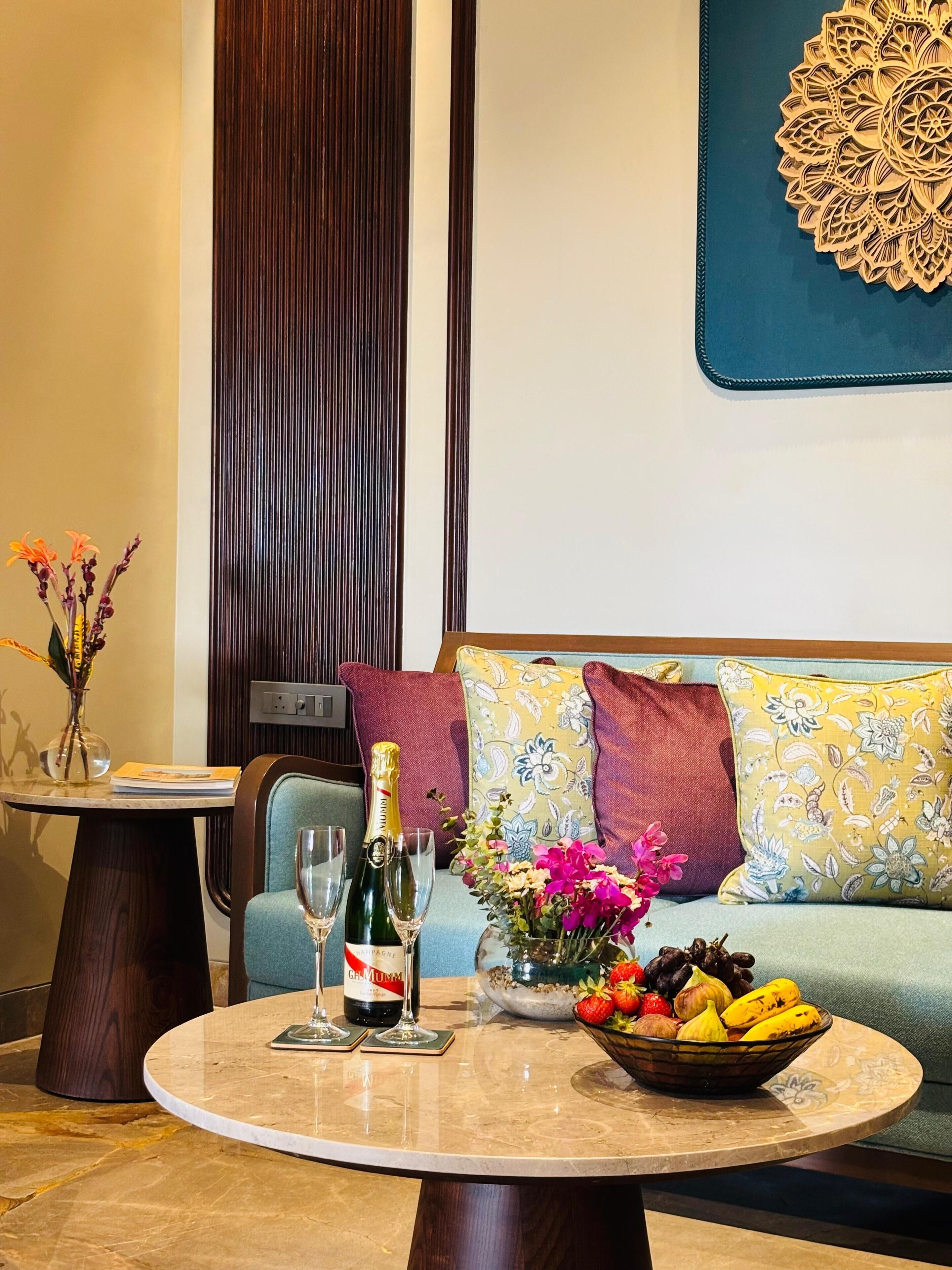 Ein Wohnzimmer mit einem runden Couchtisch, einer Champagnerflasche, zwei Weingläsern, einer Obstschale und einer Blumenvase.