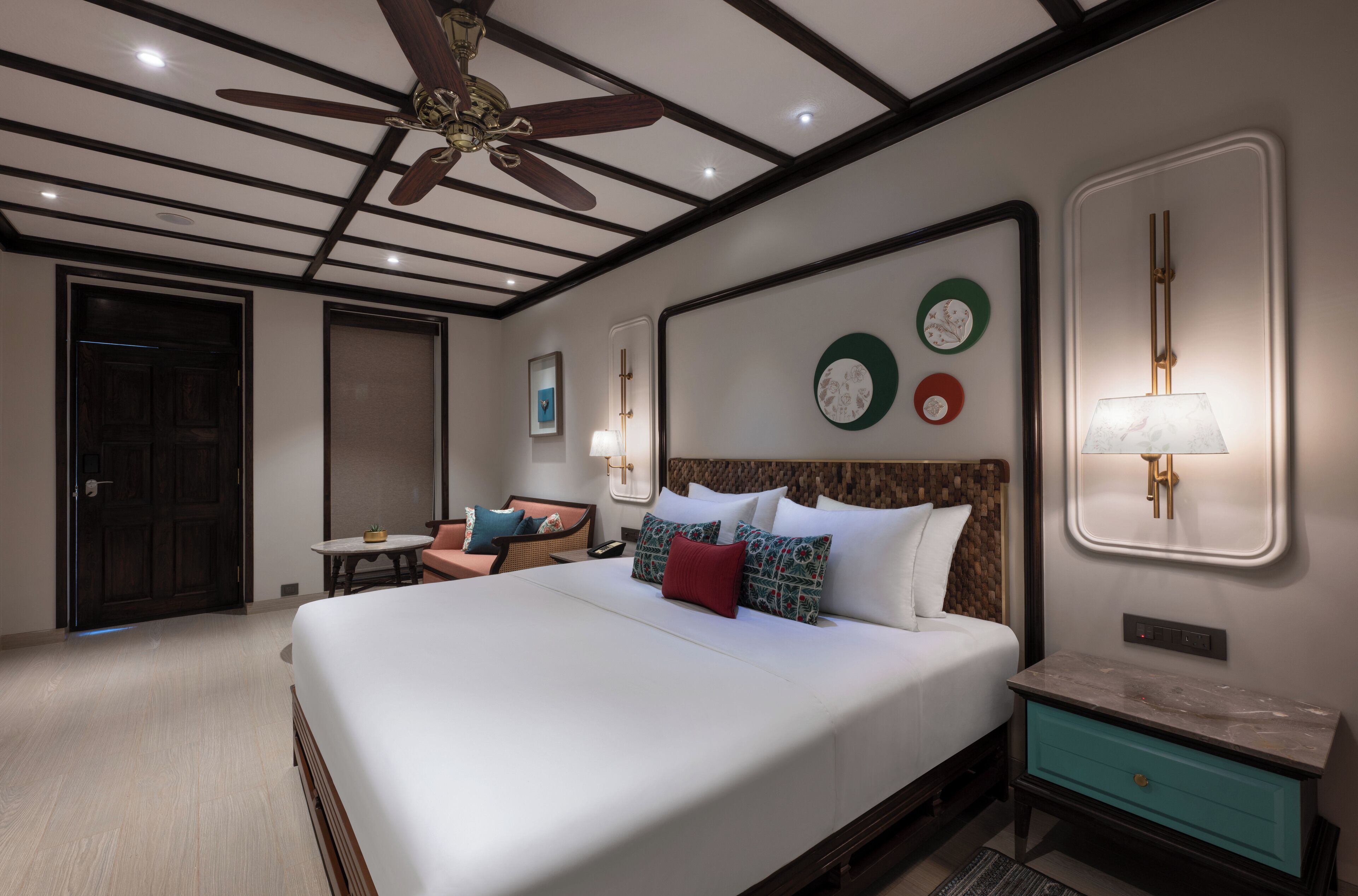 Habitación de hotel con una cama grande, ventilador de techo, una pequeña mesa con sillas, mesita de noche y relojes en la pared.
