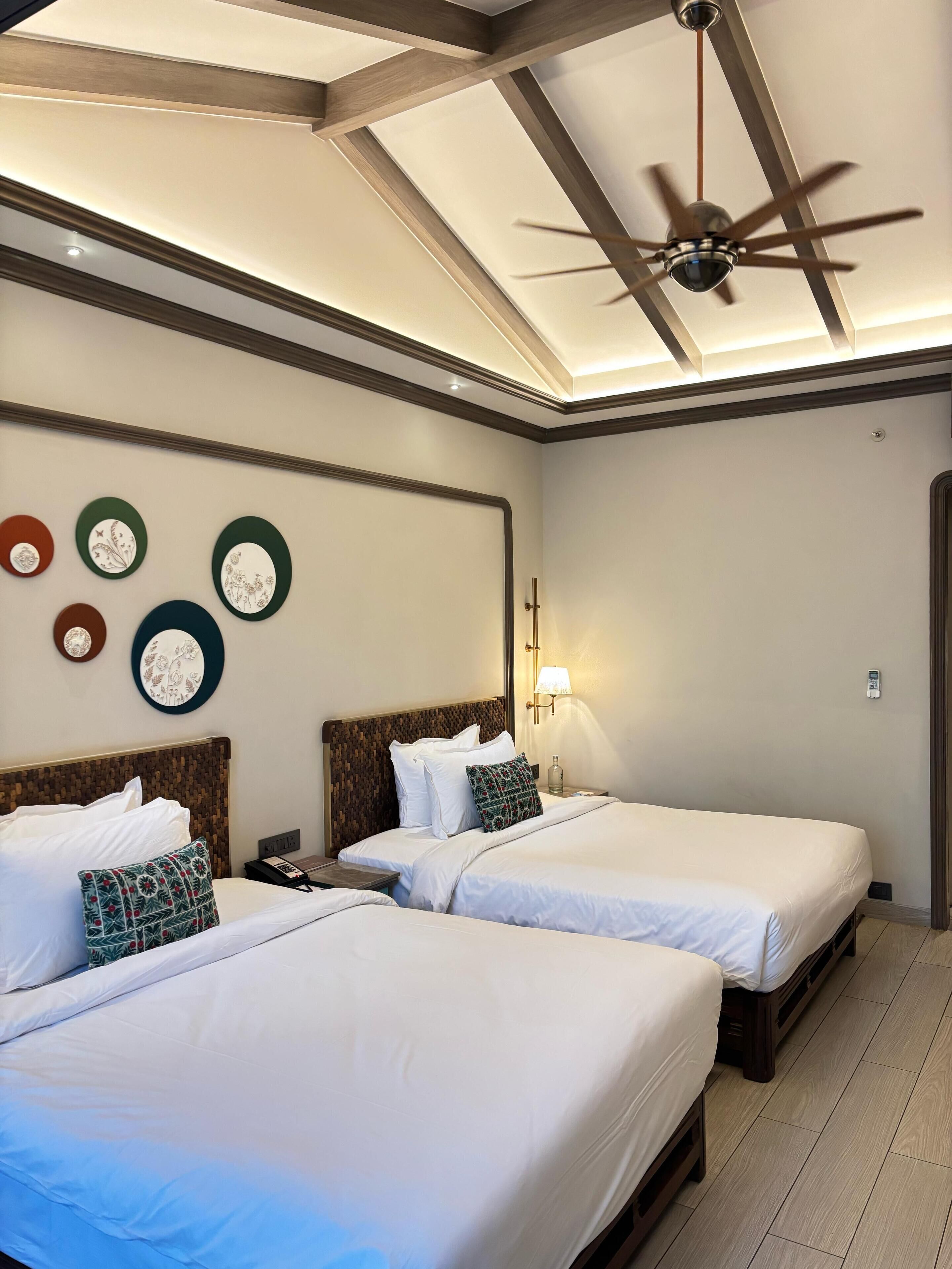 A hotel room with two beds, a ceiling fan, and wall-mounted clocks.