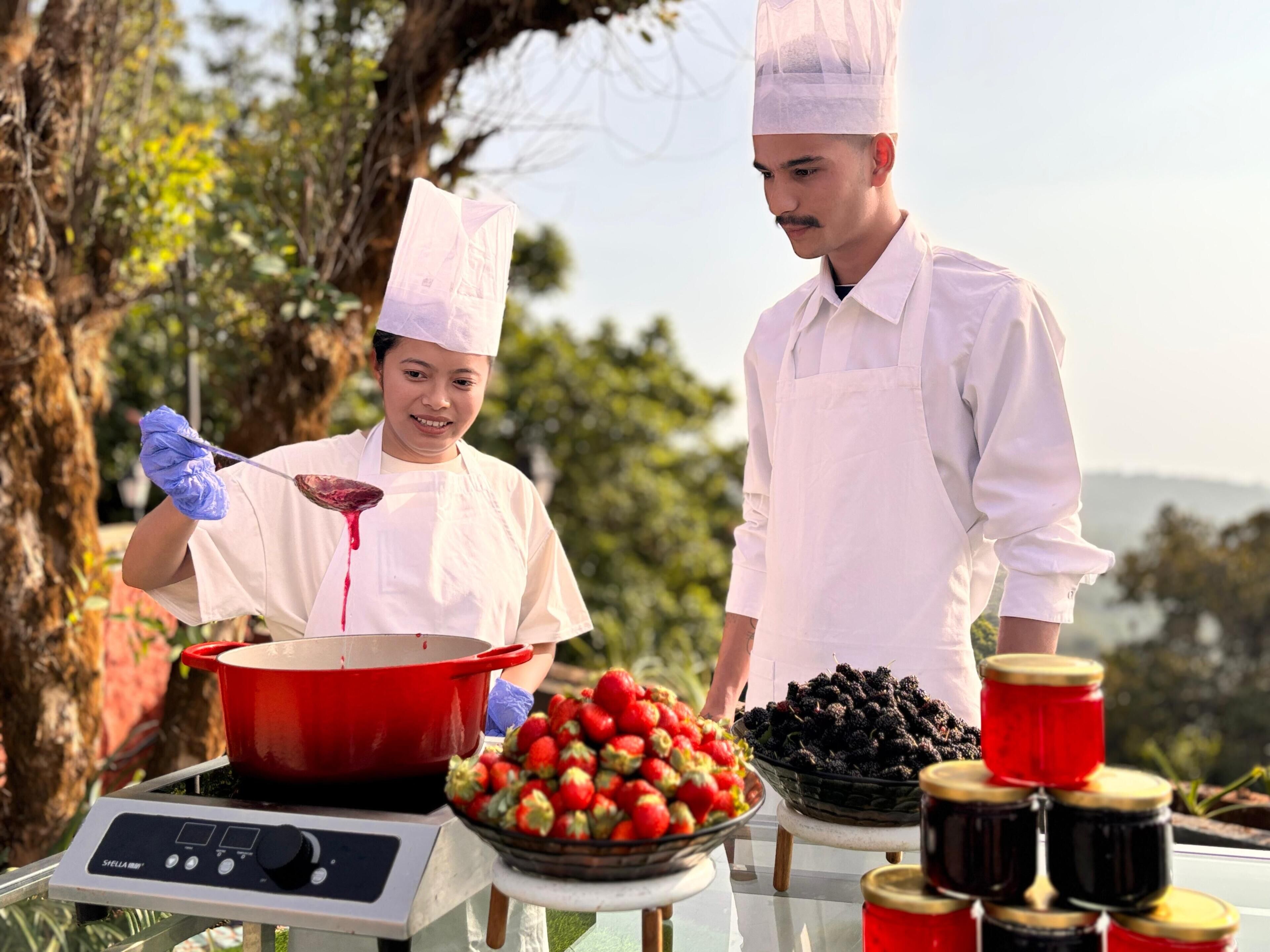 Two chefs in white uniforms and hats preparing food outdoors with a pot, strawberries, blackberries, and jars on a table.