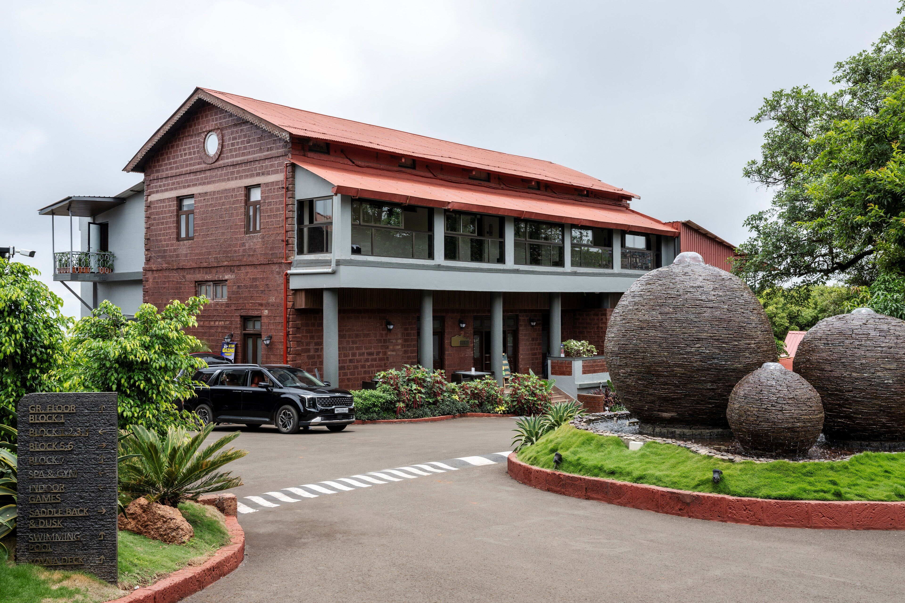 A two-story building with a red roof and a stone signboard listing amenities like a gym and pool.