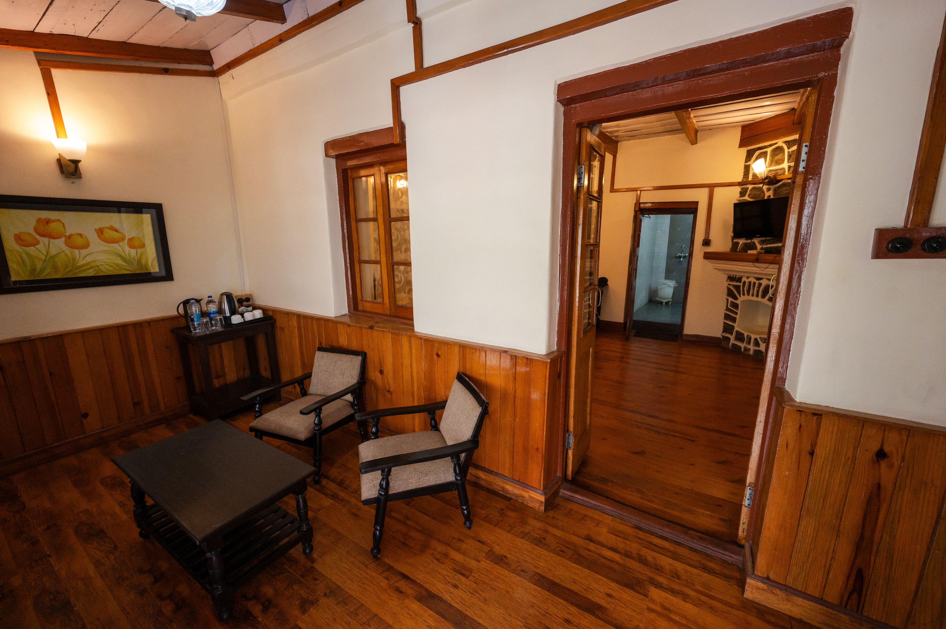 A room with wooden flooring, a table, two chairs, a painting on the wall, and a doorway leading to another room.
