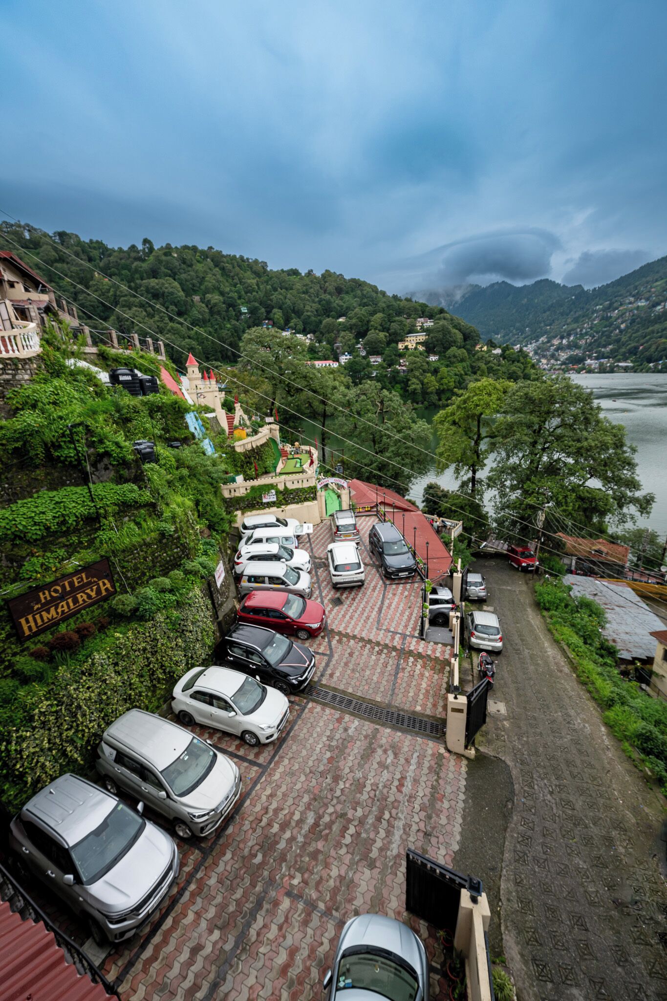 Een hotelparkeerplaats met verschillende auto's, een bord met 'Hotel Himalaya' en een uitzicht op een meer en bergen op de achtergrond.