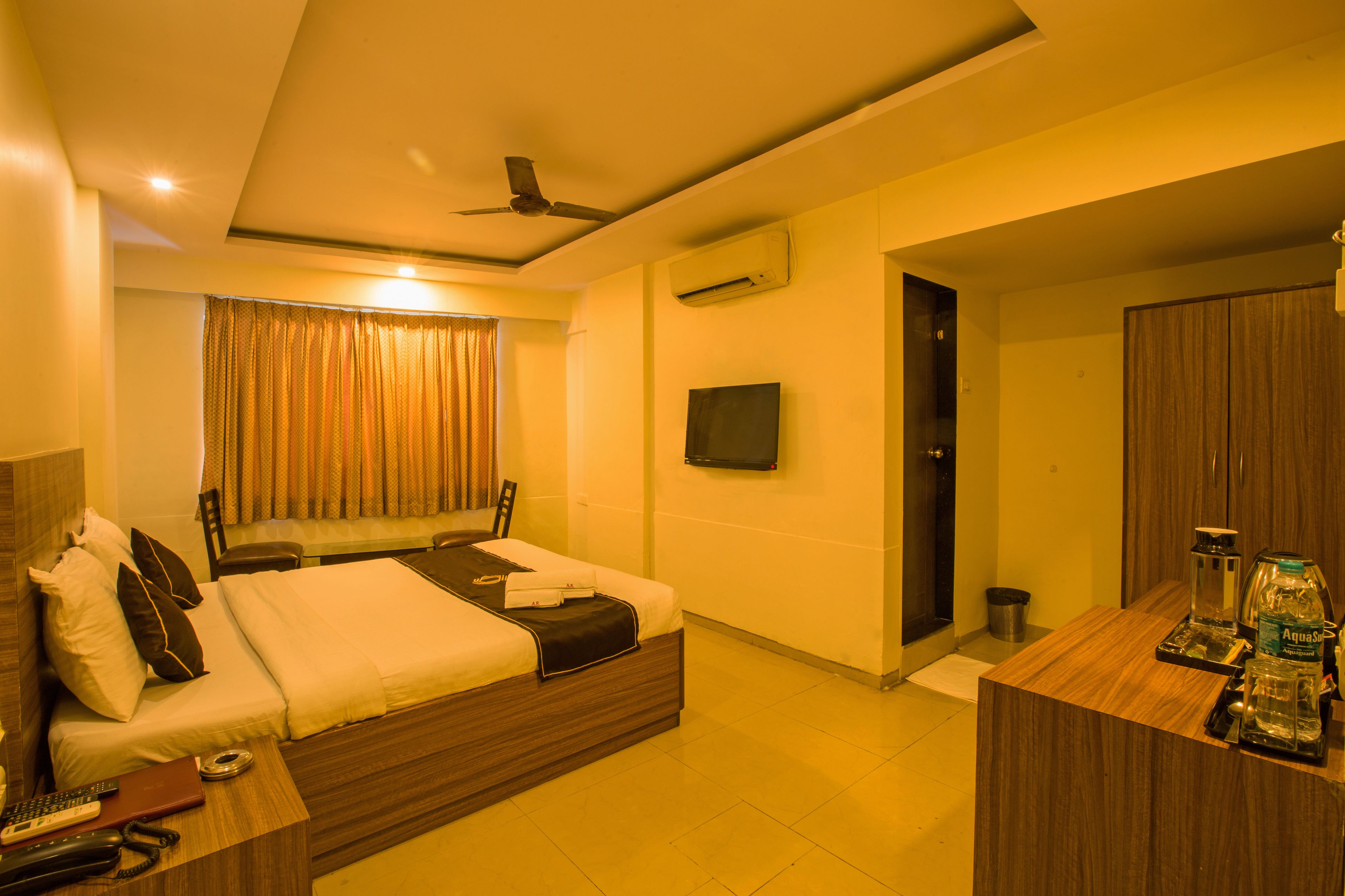 A hotel room with a bed, bedside table, TV, and a desk with a water bottle.