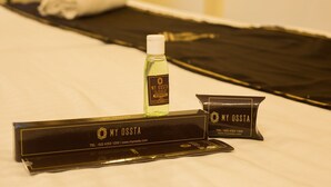 Habitación de hotel con una cama bien hecha, una botella de champú y una caja negra con el nombre y los datos de contacto del hotel.