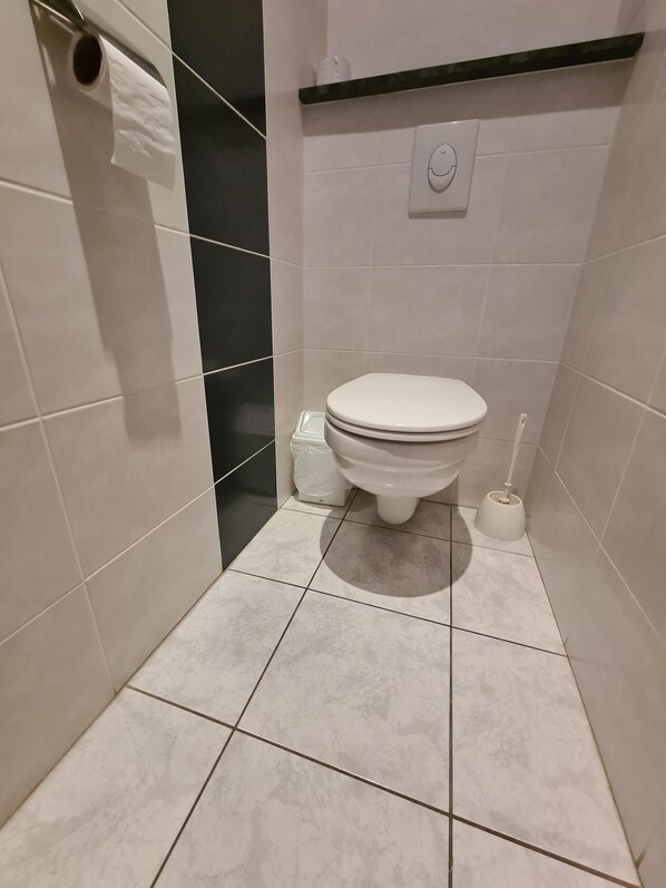 A small, clean bathroom with a toilet, a toilet brush, and a trash bin.