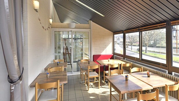 Un restaurante moderno con mesas y sillas de madera, un banco rojo y amplias ventanas que dan a la calle.